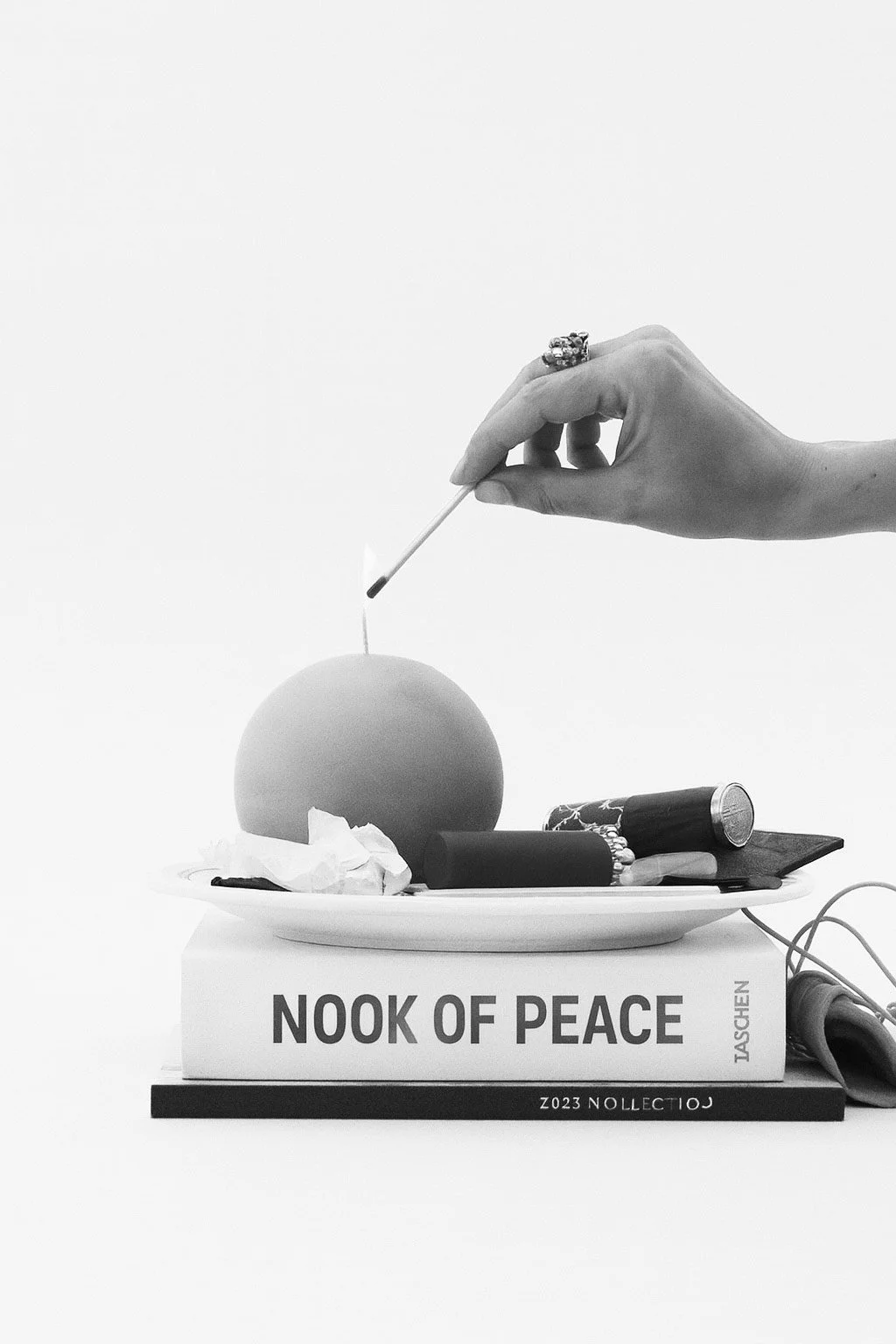 A hand holding a lit matchstick above a round object resembling an apple, placed on a plate with crumpled tissue and other objects, with books labeled "Nook of Peace" and "Taschen" underneath. The scene is in black and white.
