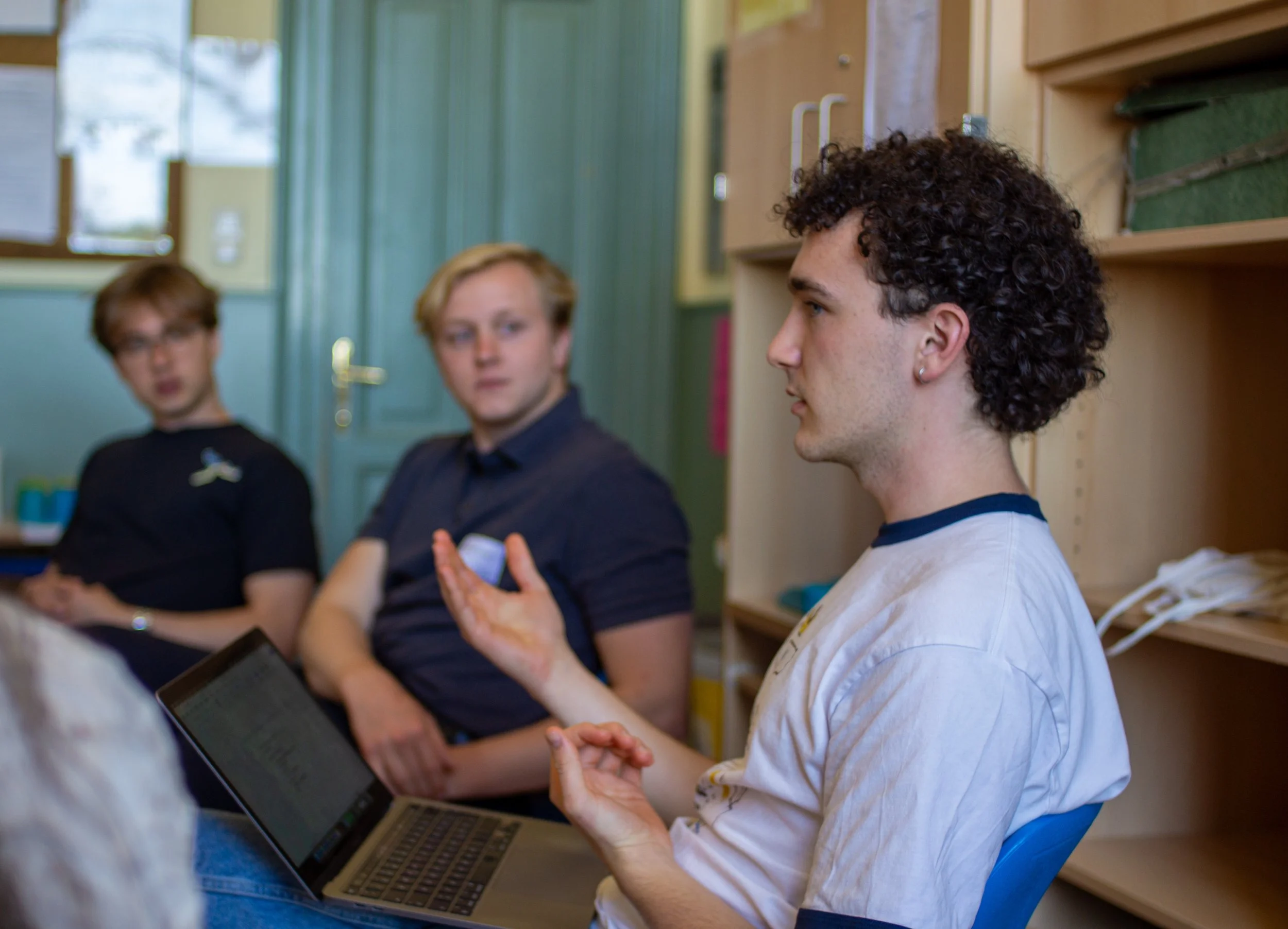 A man with curly hair, wearing a white shirt with a blue collar, sitting on a blue chair, speaking and gesturing with his hand during a discussion, with two women in the background listening attentively.