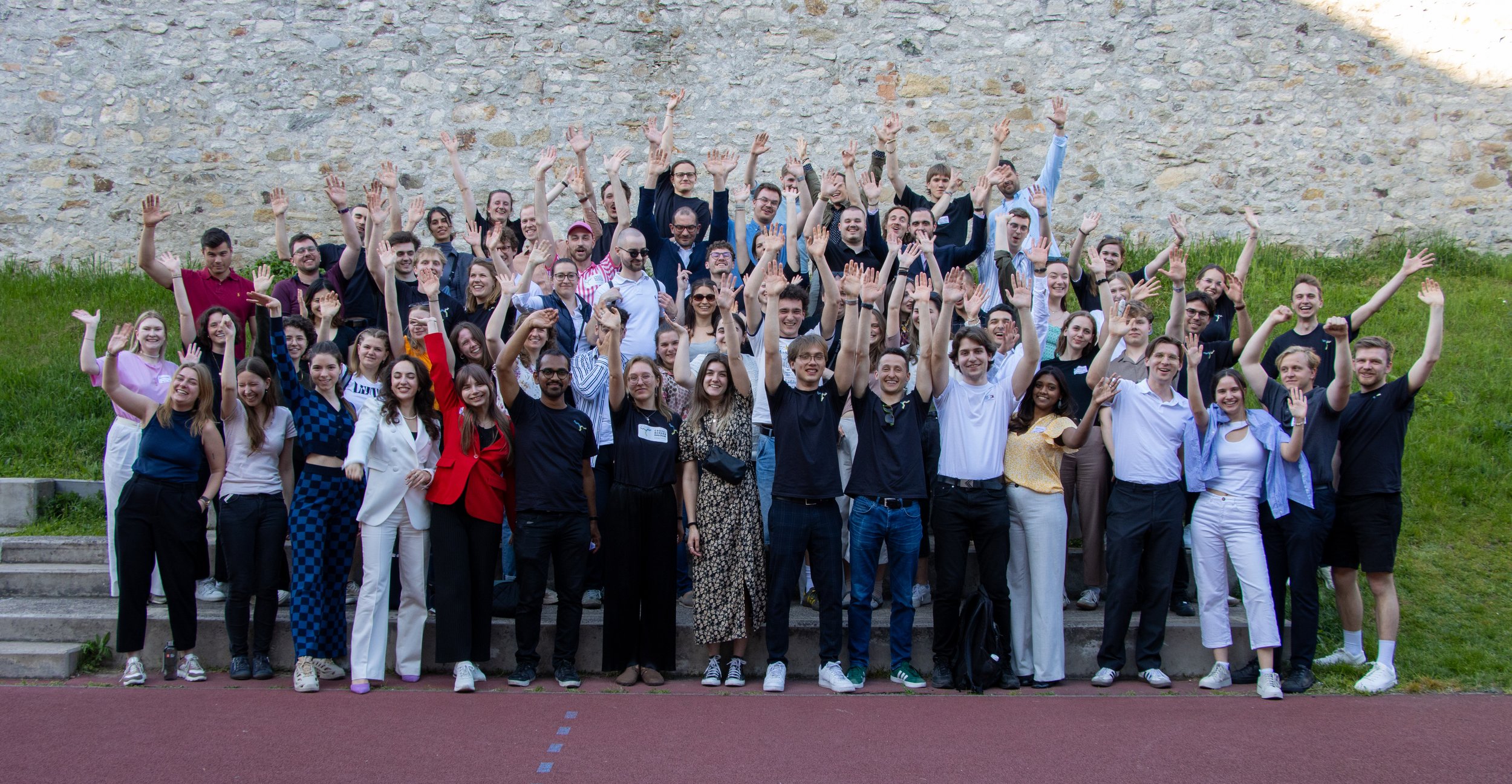 A large group of diverse young people gathered outdoors on steps, raising their hands and smiling for a group photo in front of a stone wall with grass and stairs.