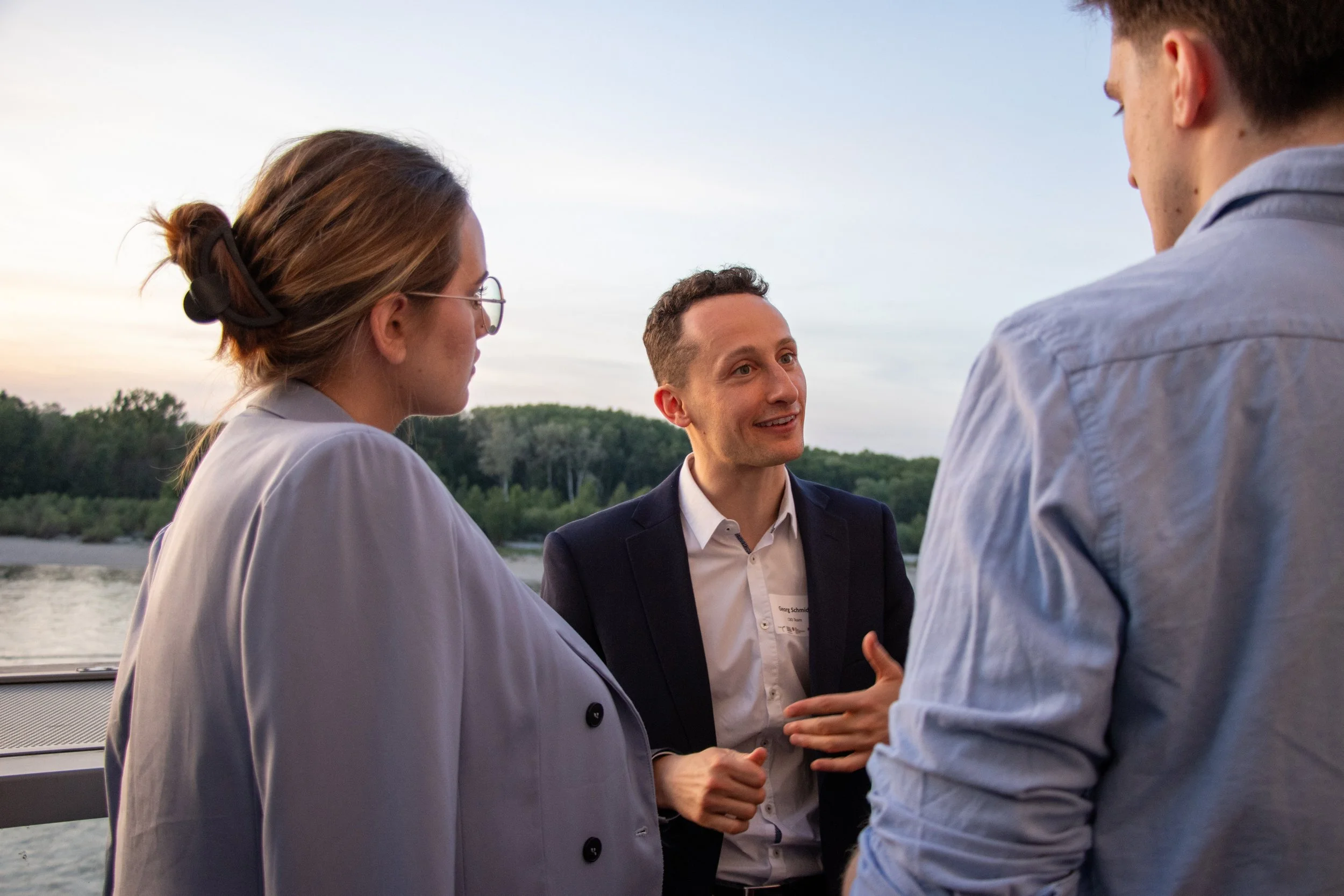 Three people are engaged in conversation outdoors near a body of water during sunset, with trees in the background. One woman has her hair tied back and is wearing glasses and a light-colored blazer. The man in the middle, wearing a dark blazer and white shirt, is speaking, while the third person has their back to the camera and is dressed in a light blue shirt.