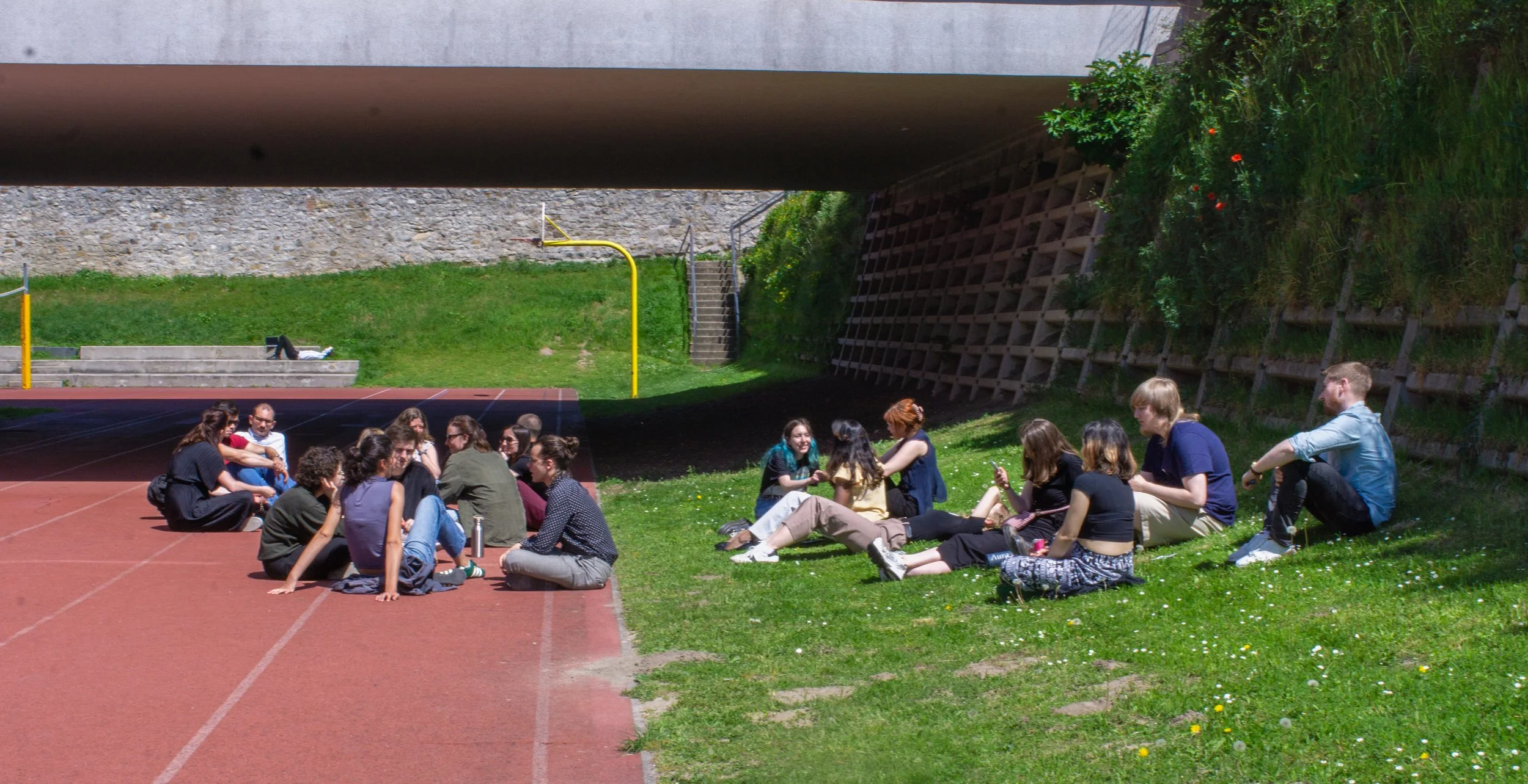 Group of people sitting and socializing on grassy area under bridge near outdoor track field.