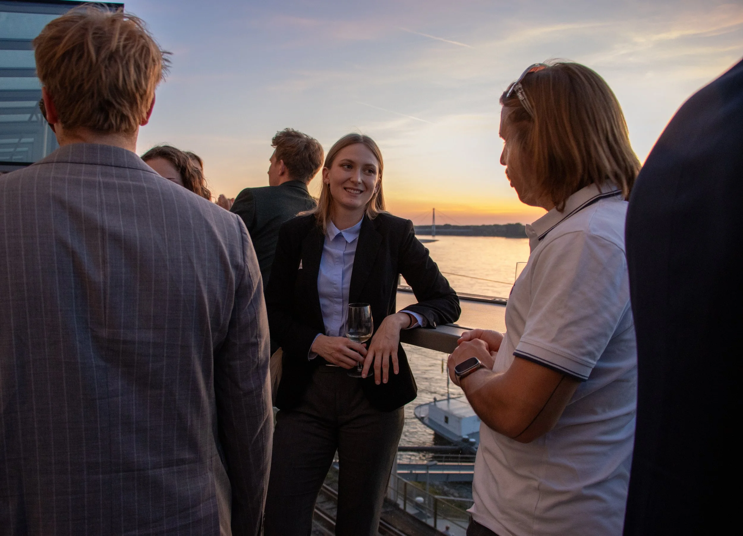 A group of people in business attire socializing outdoors on a boat deck during sunset with water and a bridge in the background.