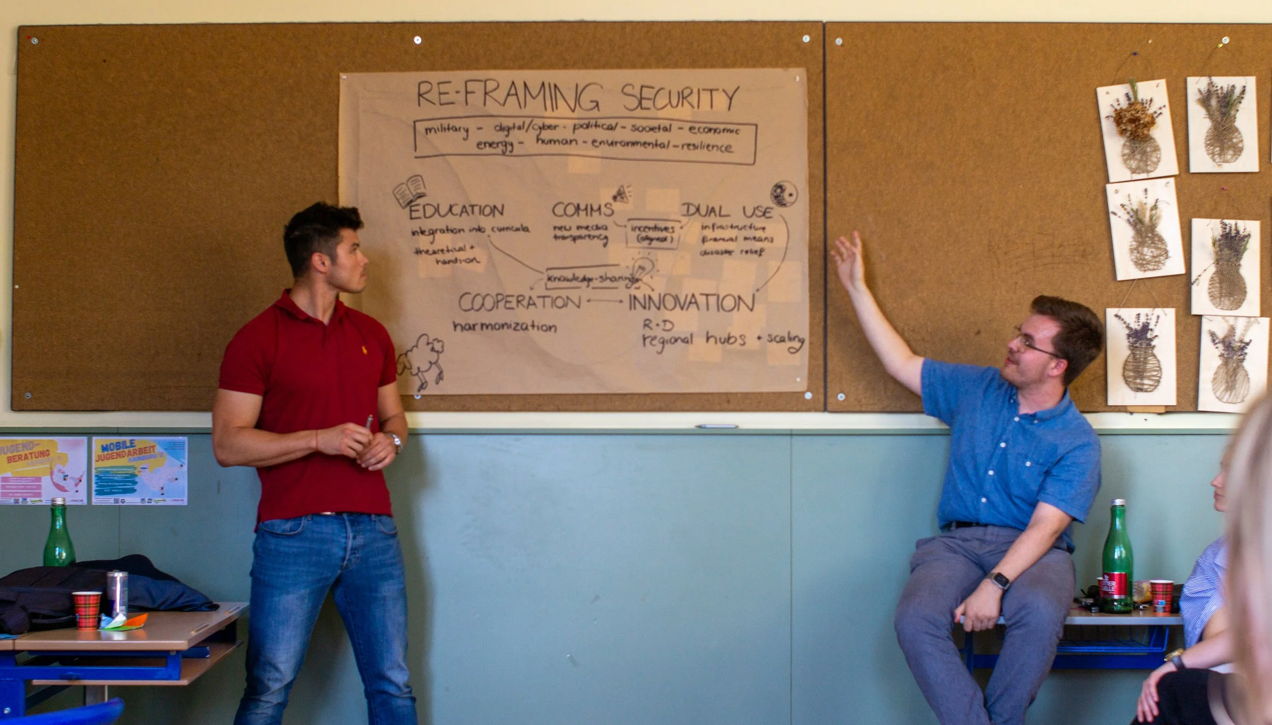 Two men are in a classroom, one standing and the other sitting on a small table, engaged in discussion about a large poster titled 'Re- framing Security' on a cork board. The poster contains handwritten notes and diagrams related to security.