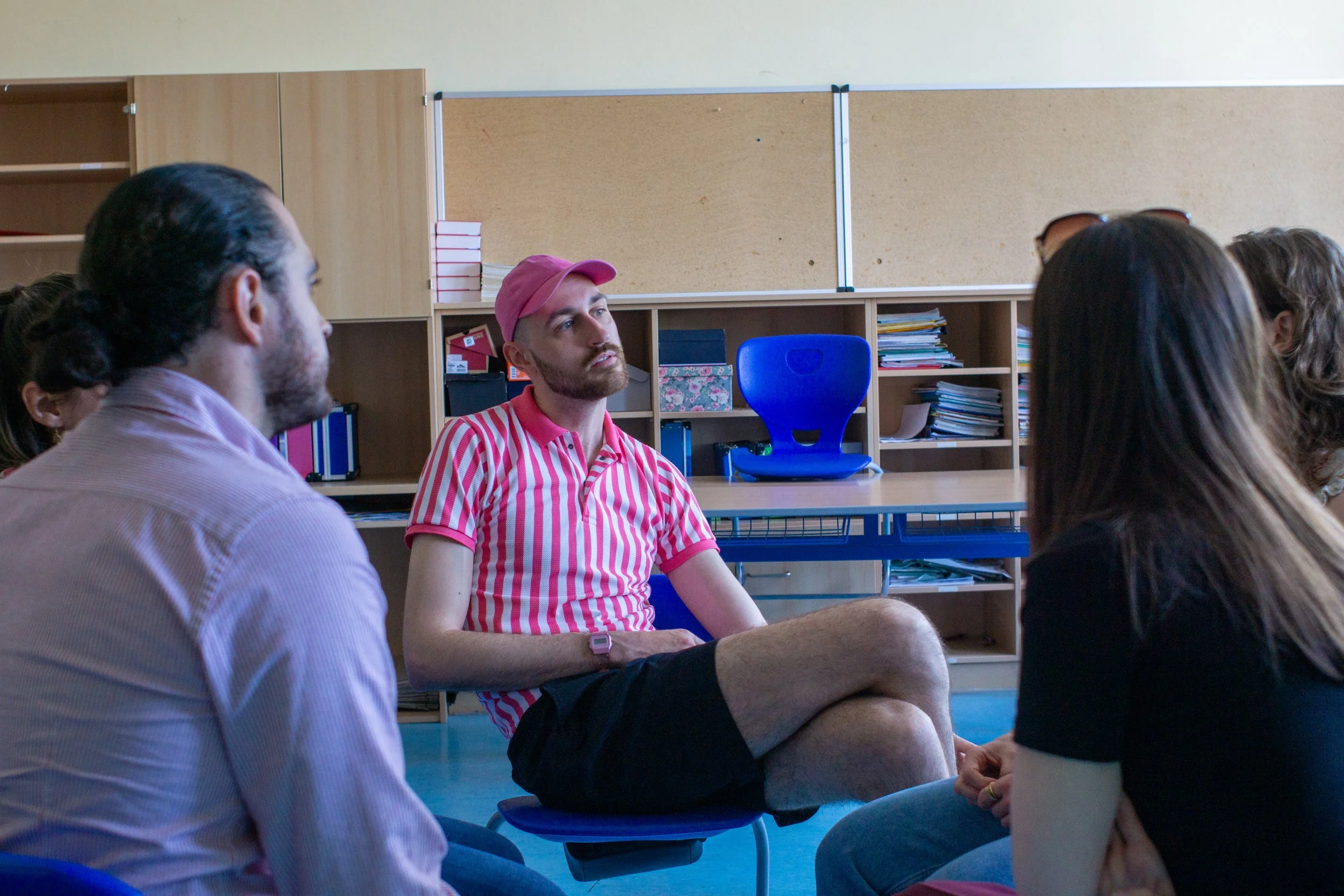 A group of people sitting in a circle in a classroom or meeting room. A man in a pink and white striped shirt and pink cap is speaking. The others are listening attentively.