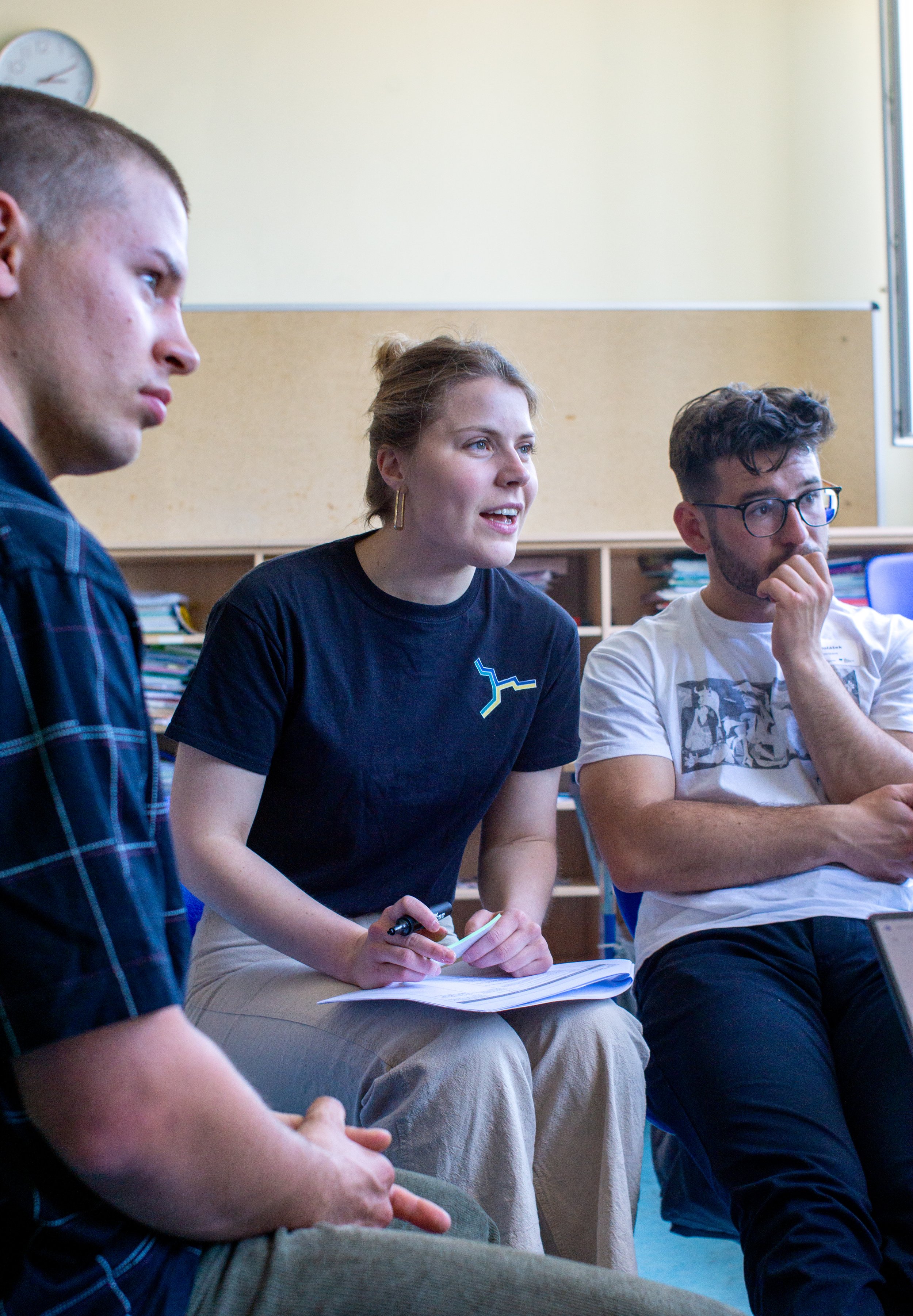 Three young adults sitting in a classroom, engaged in a discussion. One woman in the center is speaking, holding a pen and notepad, while a man with glasses on the right listens thoughtfully. Another person is partially visible on the left.