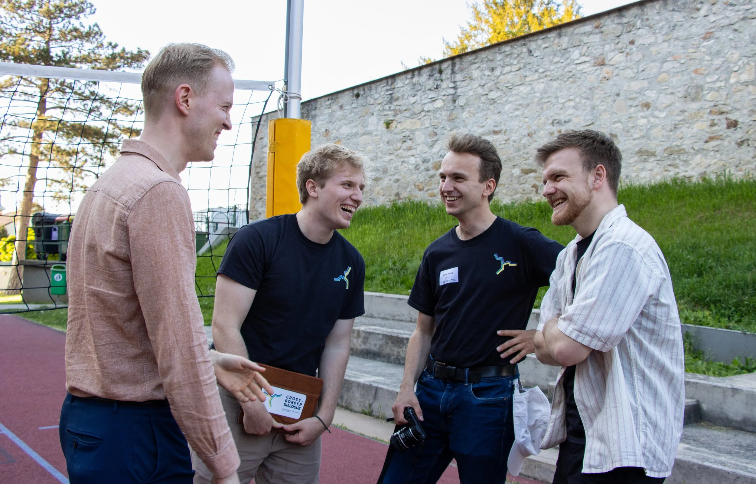 Four men standing outdoors by a volleyball net, engaged in conversation and smiling. Two are wearing black t-shirts with a logo, one in a pink shirt, others in casual shirts.