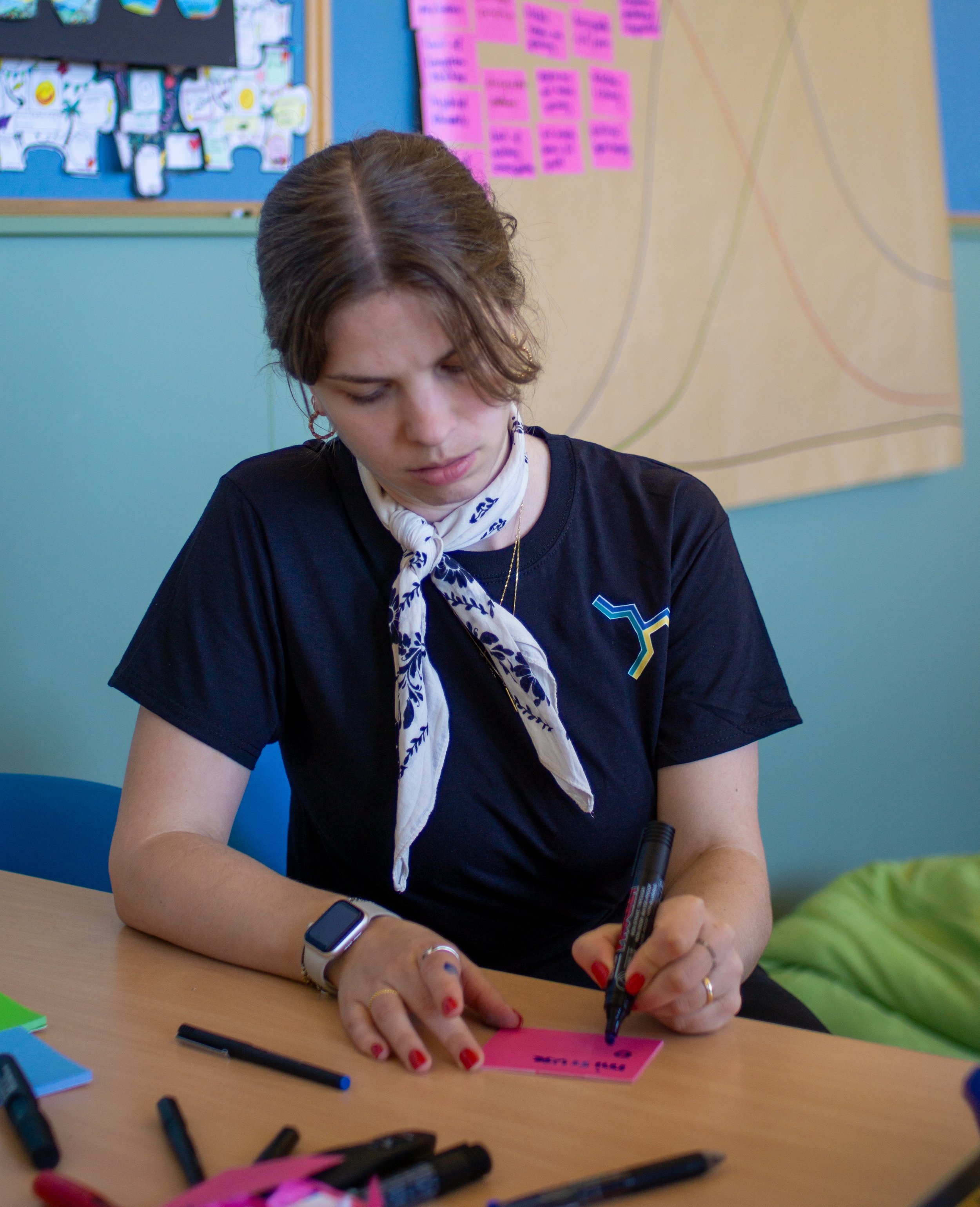 A woman with brown hair, wearing a black shirt, white scarf, and smartwatch, sitting at a table and writing on a pink sticky note with a black marker in a classroom setting.