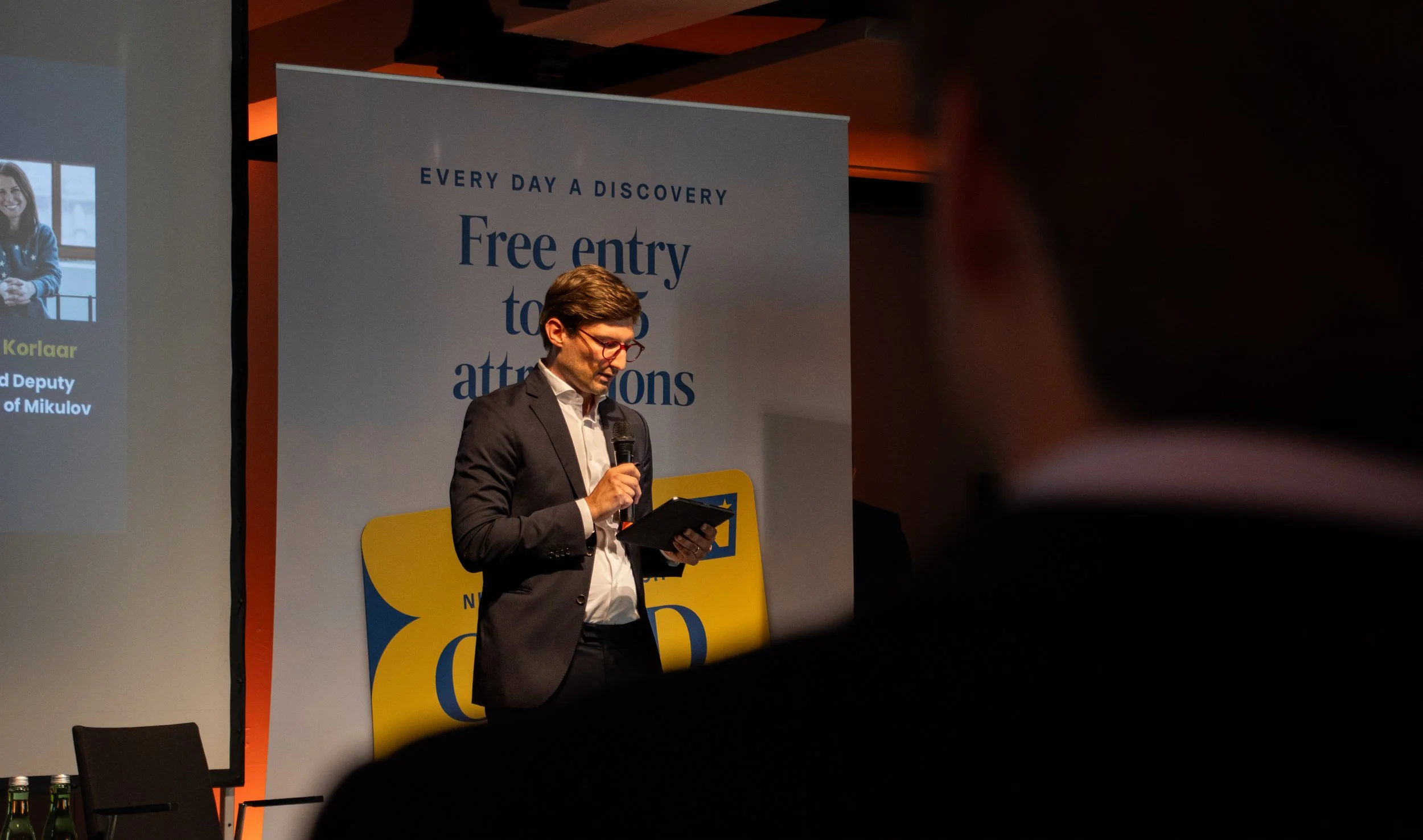 A man in a dark suit with red glasses holds a microphone and reads from a tablet on stage at an event. Behind him is a large white banner with blue text that reads 'Every day a discovery, Free entry to attractions.' To the left, a partly visible screen shows a woman in a blue jacket, and some green bottles are on a table.