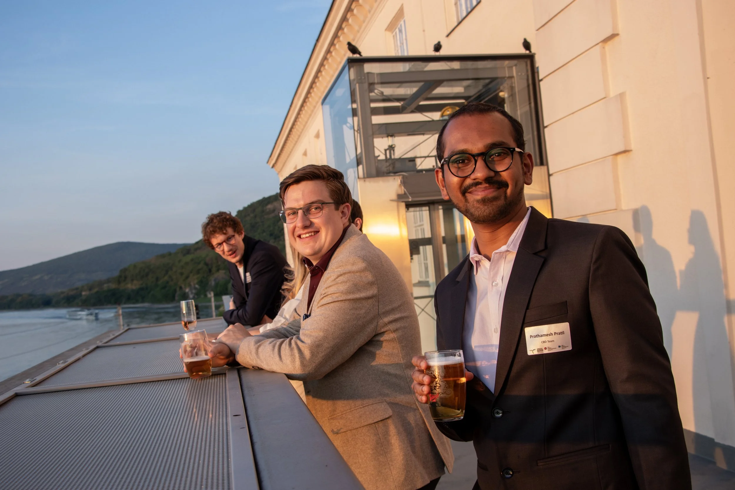 Four people in business attire enjoying drinks on a rooftop balcony overlooking water and mountains at sunset.