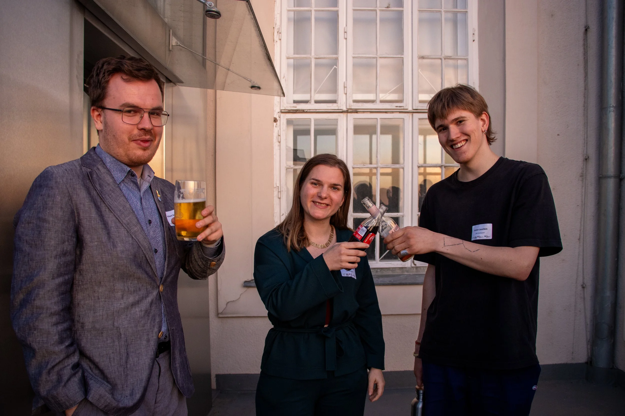 Three people smiling on a rooftop, two men and one woman, holding drinks in their hands.