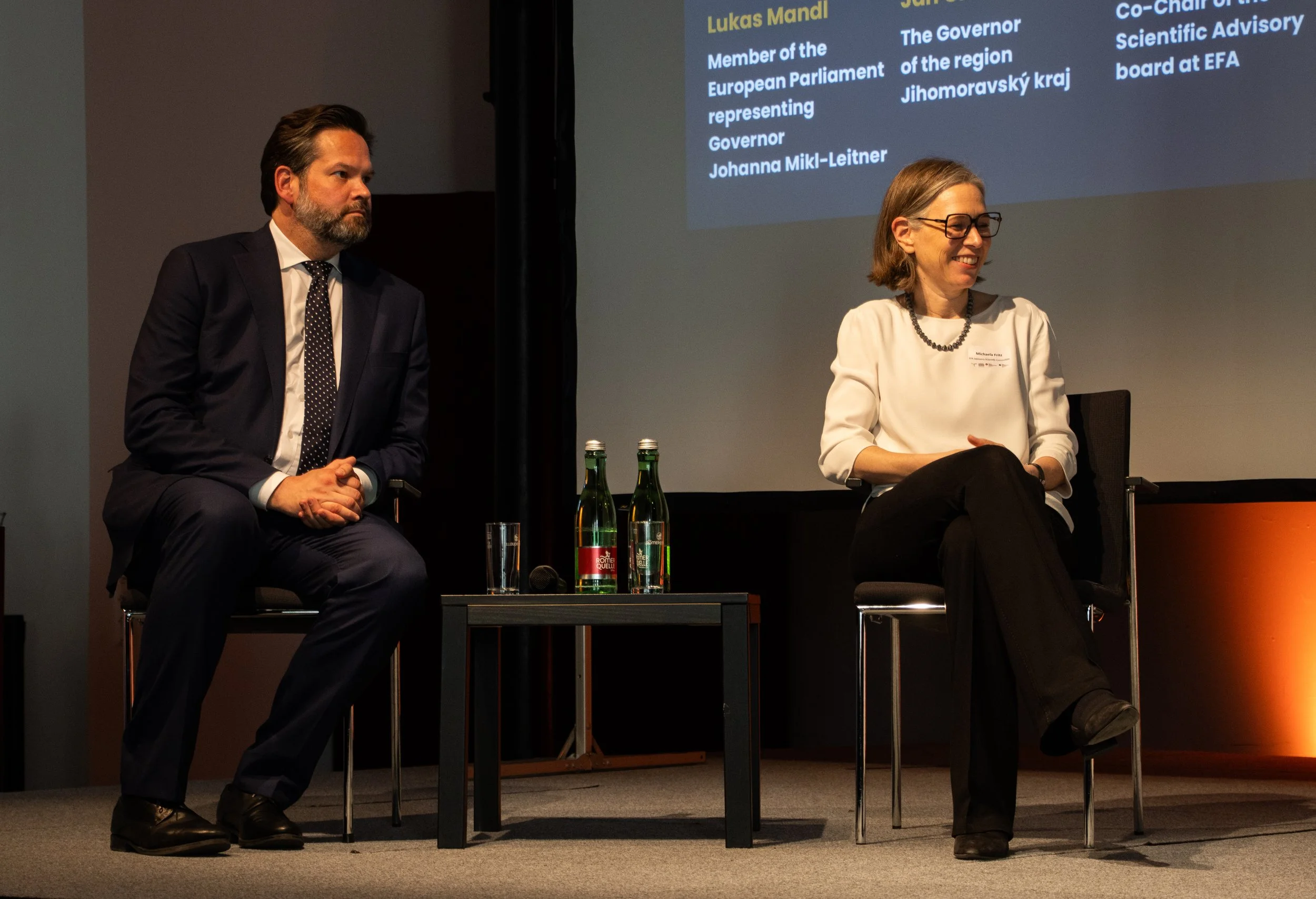 Two people sitting on chairs in front of a presentation screen, a man on the left in a navy suit and a woman on the right in a white blouse with a necklace, both engaged in a panel discussion. There are two bottles of water and a glass on a small table between them.