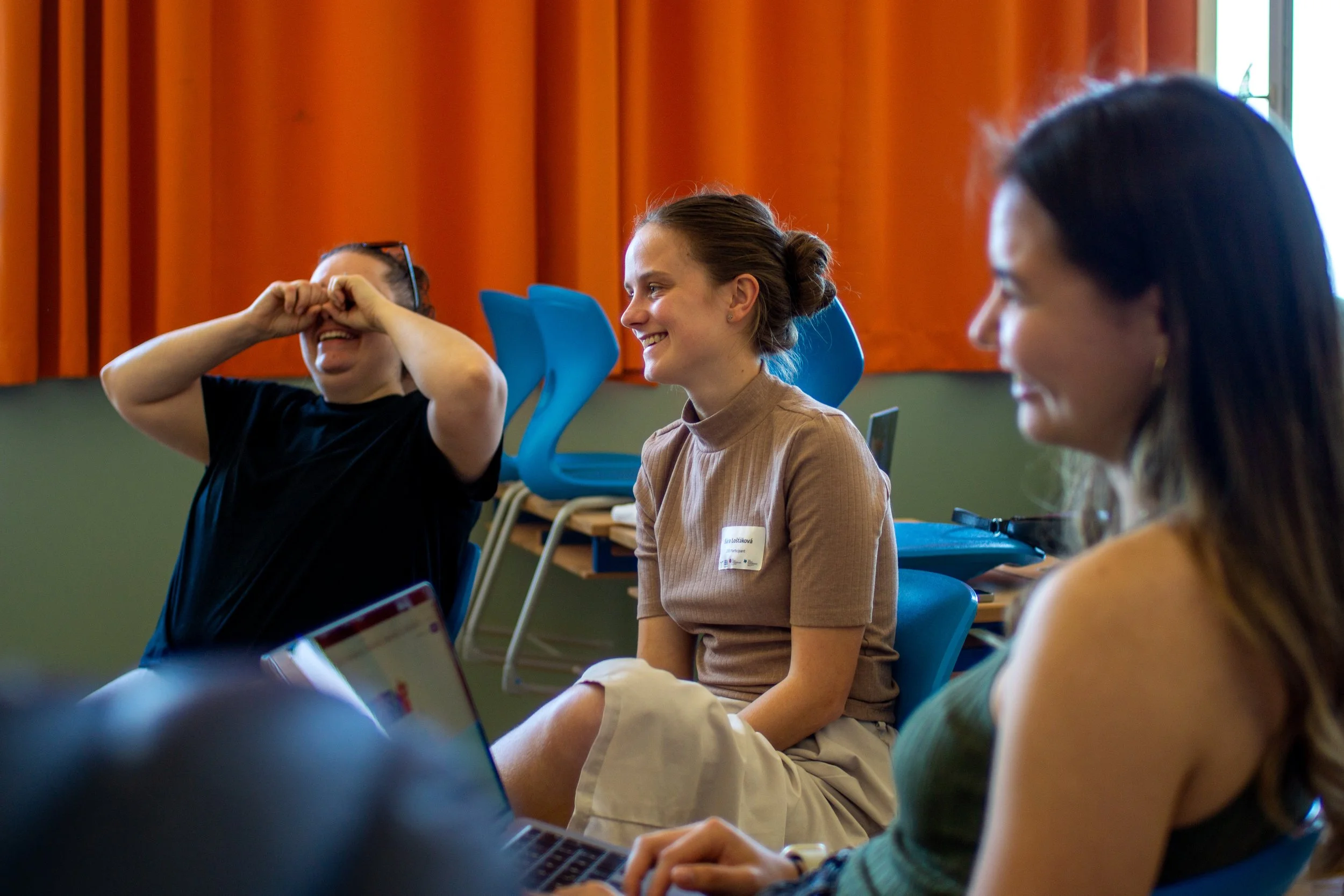 Three women sitting together in a room with orange curtains, smiling and laughing during a discussion or meeting.