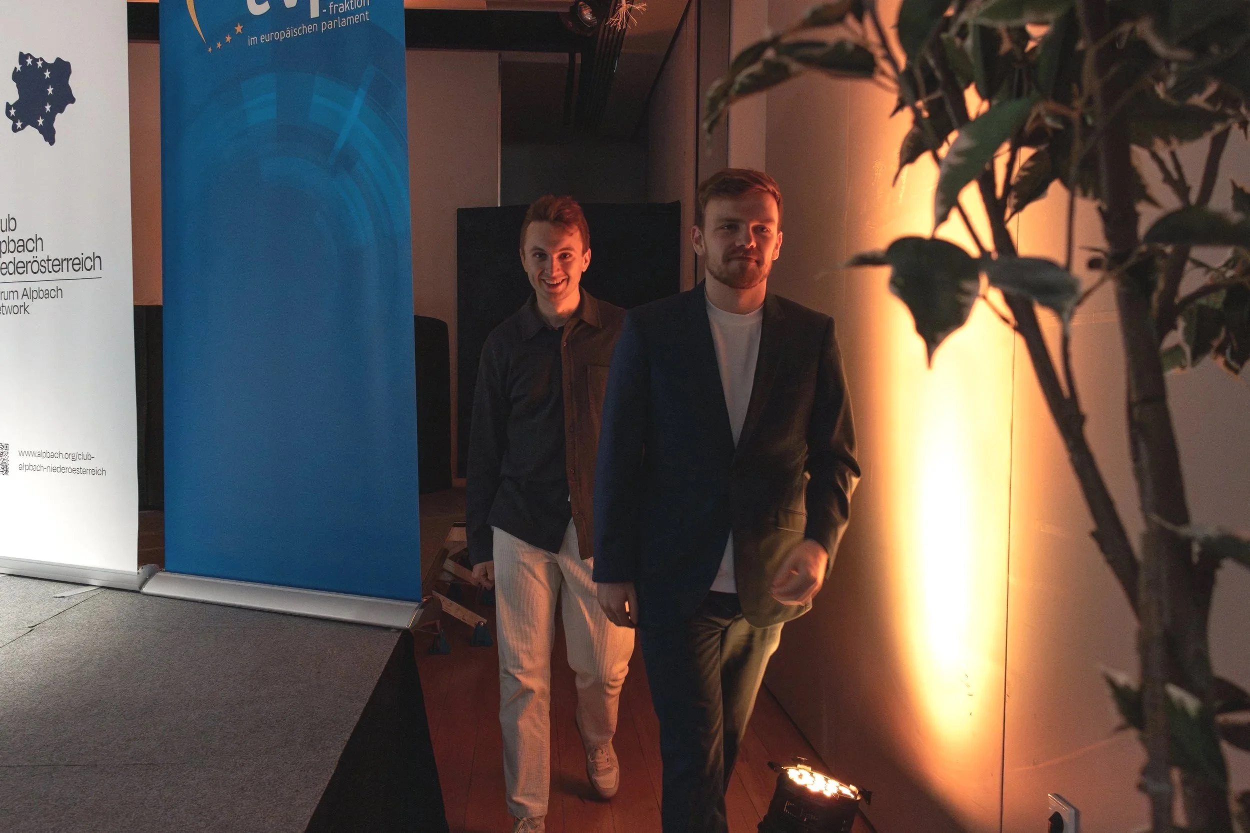 Two young men walking in a dimly lit indoor event space, one smiling and wearing a dark shirt, the other with a serious expression in a dark suit, with banners in the background and a potted plant on the right.