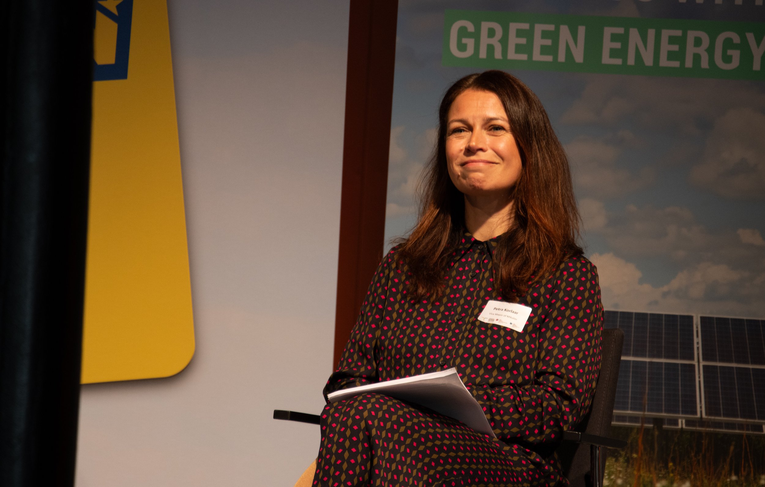 A woman with long brown hair smiling and sitting on a stage, wearing a black dress with red and pink polka dots, holding a notebook in her lap, with a backdrop showing solar panels and cloudy sky, and a banner that reads 'GREEN ENERGY'.