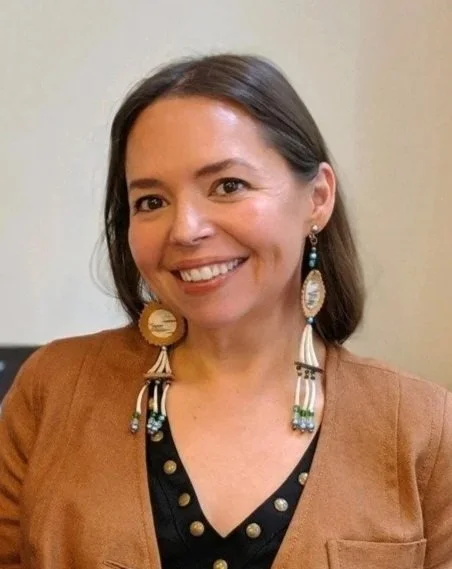 A woman with shoulder-length dark hair smiling, wearing large feather earrings, a brown blazer, and a black top with gold buttons, standing indoors against a plain background.