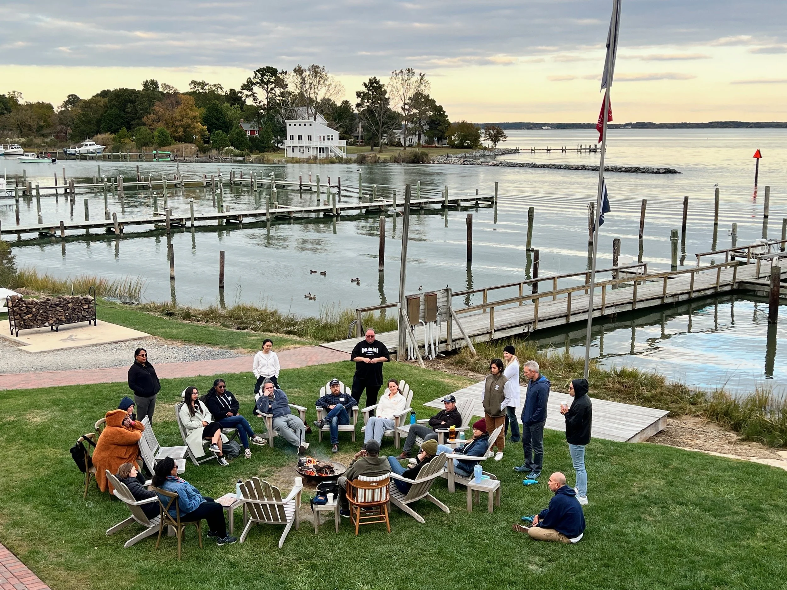 A group of people gathered outdoors near a oceanside, seated on lawn chairs and standing, with a wooden dock extending into the water and ducks swimming nearby, during twilight.