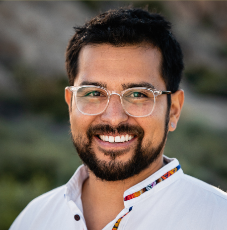 Portrait of a smiling man with glasses, short dark hair, and a beard, wearing a white shirt with a colorful collar, outdoors with a blurred natural background.