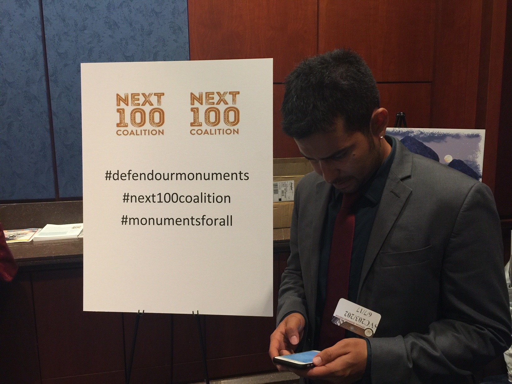 A man in a suit looking at his phone next to a sign that reads 'Next 100 Coalition' with hashtags #defourdmonuments, #next100coalition, and #monumentsforall.