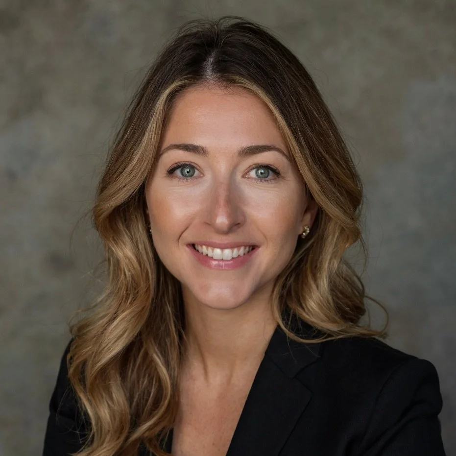 A professional woman with long wavy hair and blue eyes, wearing a black blazer, smiling at the camera against a gray background.