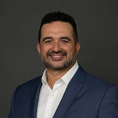 Headshot of a smiling man with dark hair and a beard, wearing a navy blazer and white shirt, against a gray background.