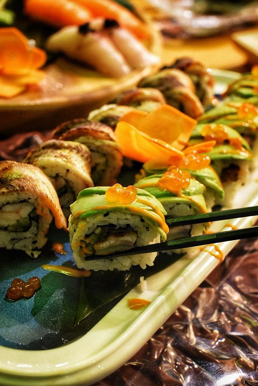Close-up of assorted sushi rolls with avocado, fish, and sauce, on a tray with chopsticks.