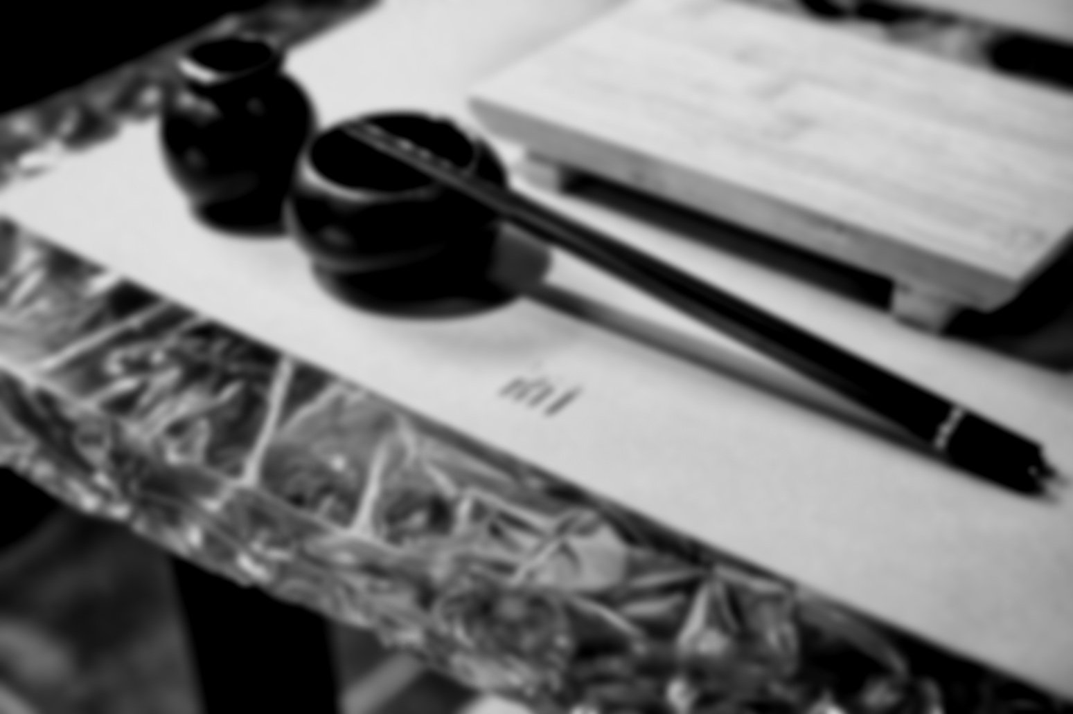 Black and white photo of a desk with a pen, stacks of paper, headphones, and a closed book on top, near a brick wall.