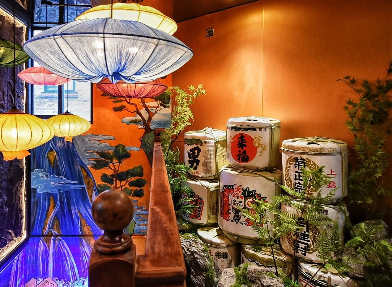 Decorative scene with Japanese sake barrels stacked next to a painted wall with traditional Japanese art, surrounded by plants and illuminated colorful paper lanterns.