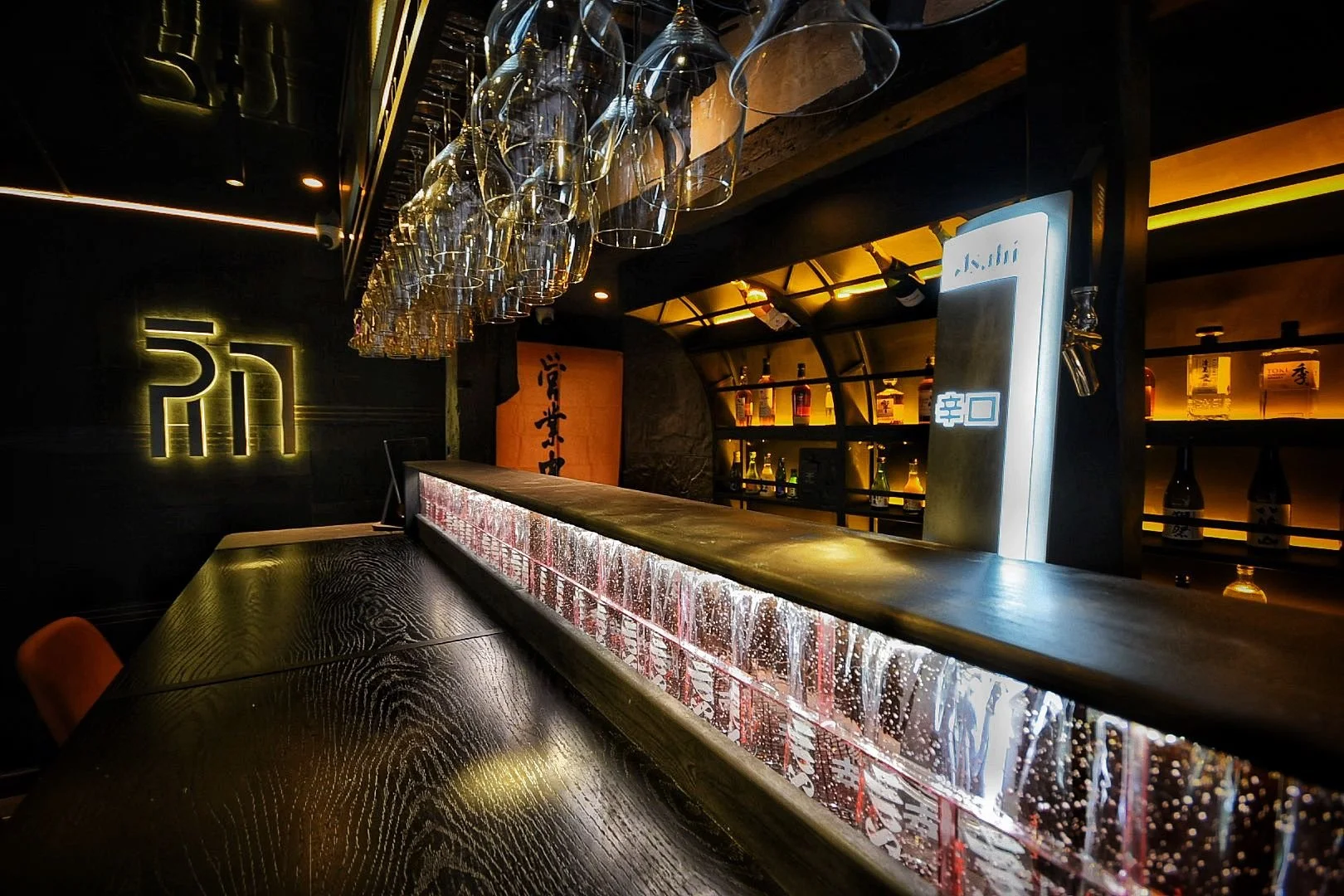 A modern, dimly-lit bar with hanging wine glasses above a wooden counter. Behind the bar, shelves with bottles of liquor are illuminated with warm yellow lighting. A neon sign with Asian characters is on the left wall.