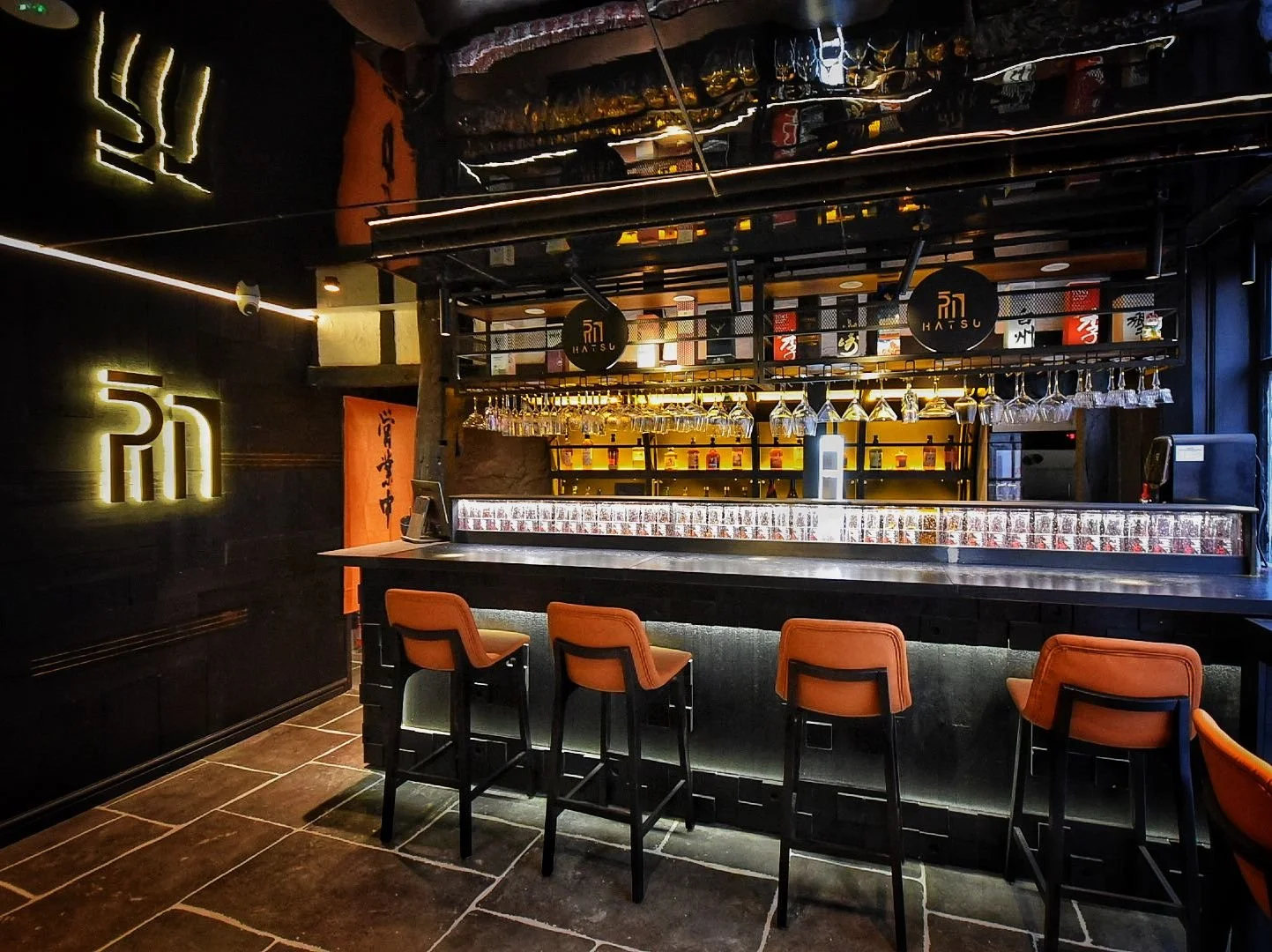 Interior of a modern Japanese-style bar with a black wall, illuminated matching signage, and a high counter with four orange chairs. The bar has hanging wine glasses, shelves with liquor bottles, and a Zen-inspired decor with neon accents.