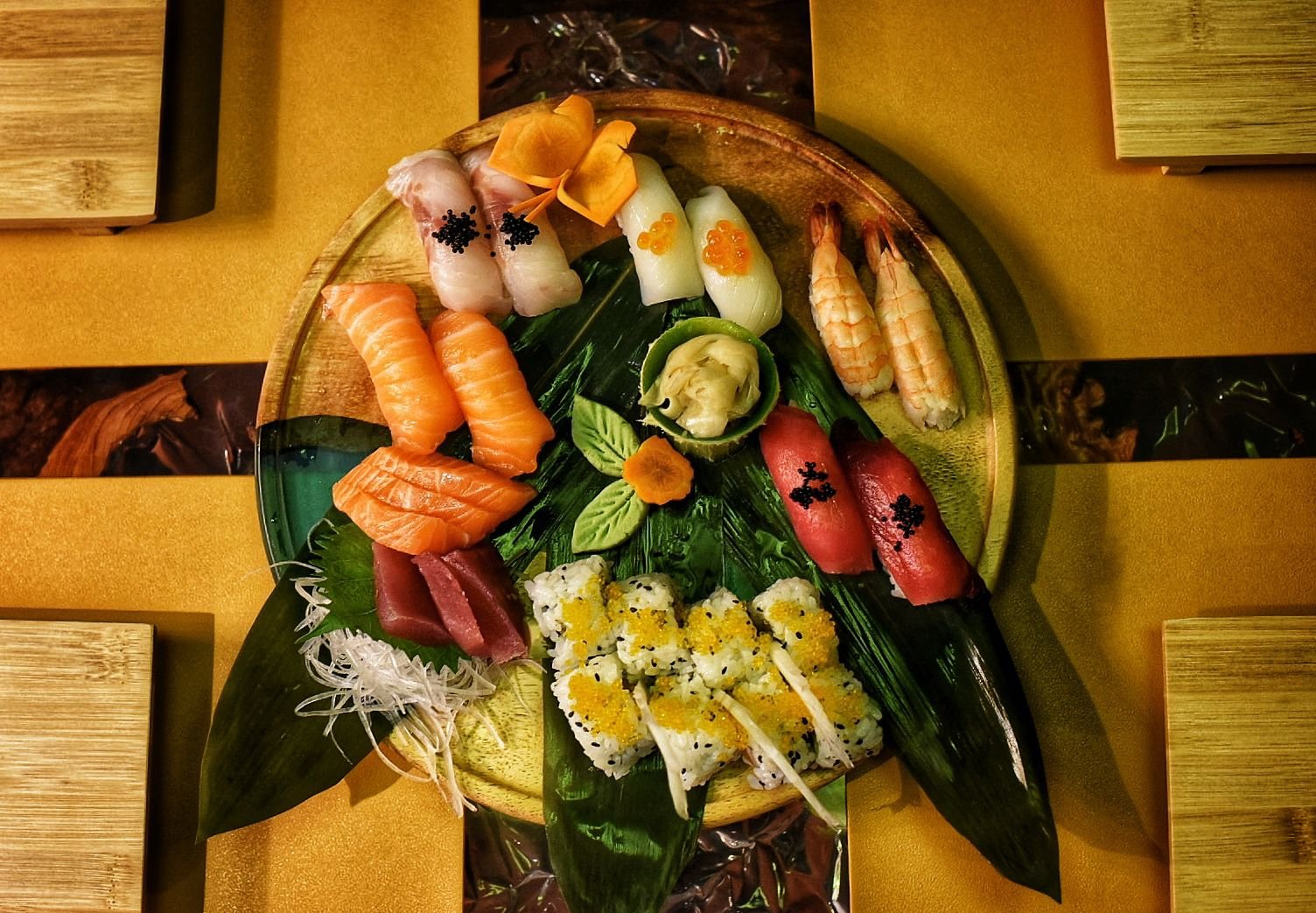 Assorted sushi platter with various types of sushi including salmon, tuna, shrimp, and nigiri, garnished with leaves and decorative vegetables, on a round wooden plate.