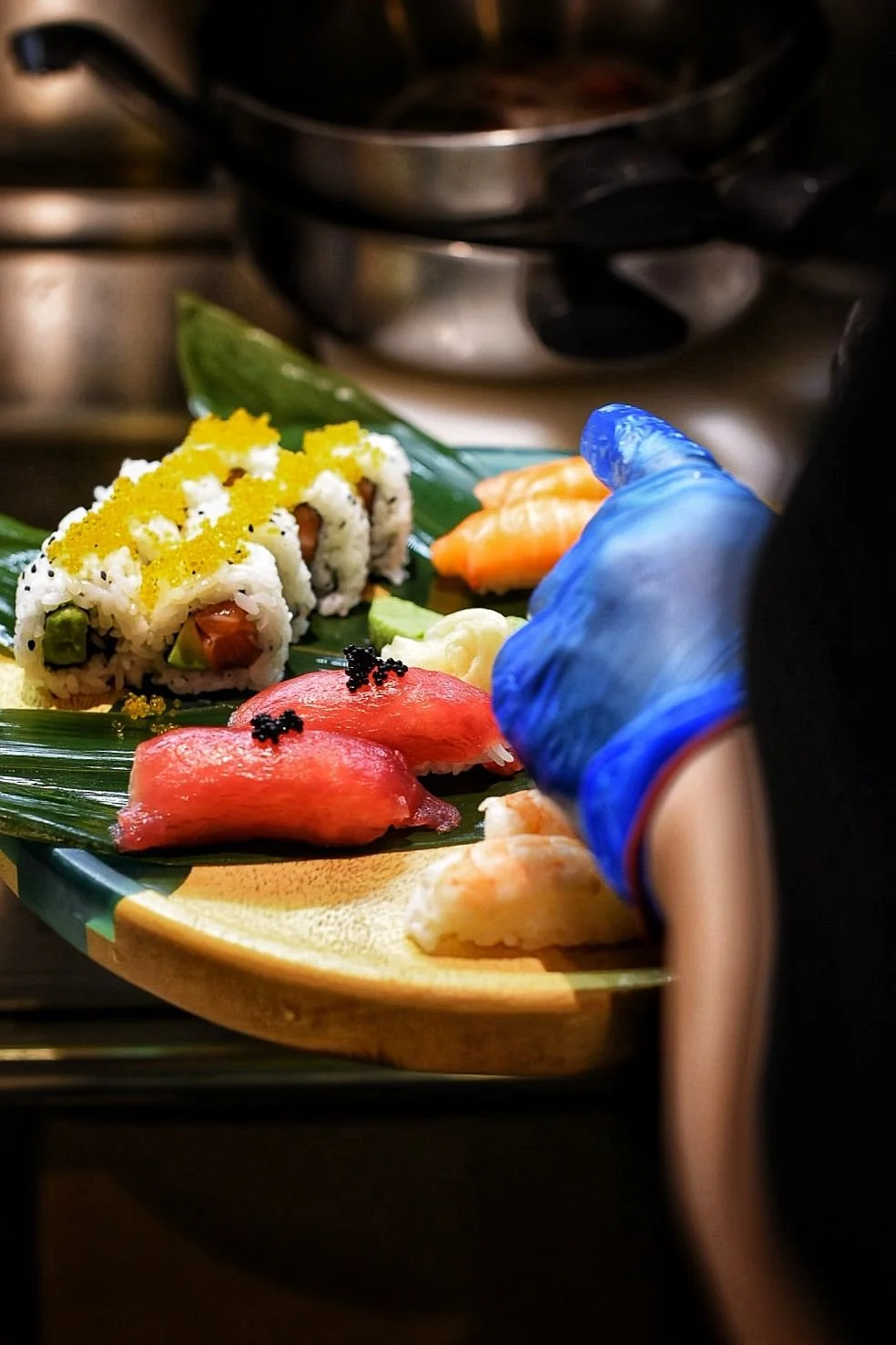 A chef's hand, wearing a blue glove, prepares assorted sushi on a wooden platter. The sushi includes slices of raw fish, sushi rolls with various fillings, and garnishes.