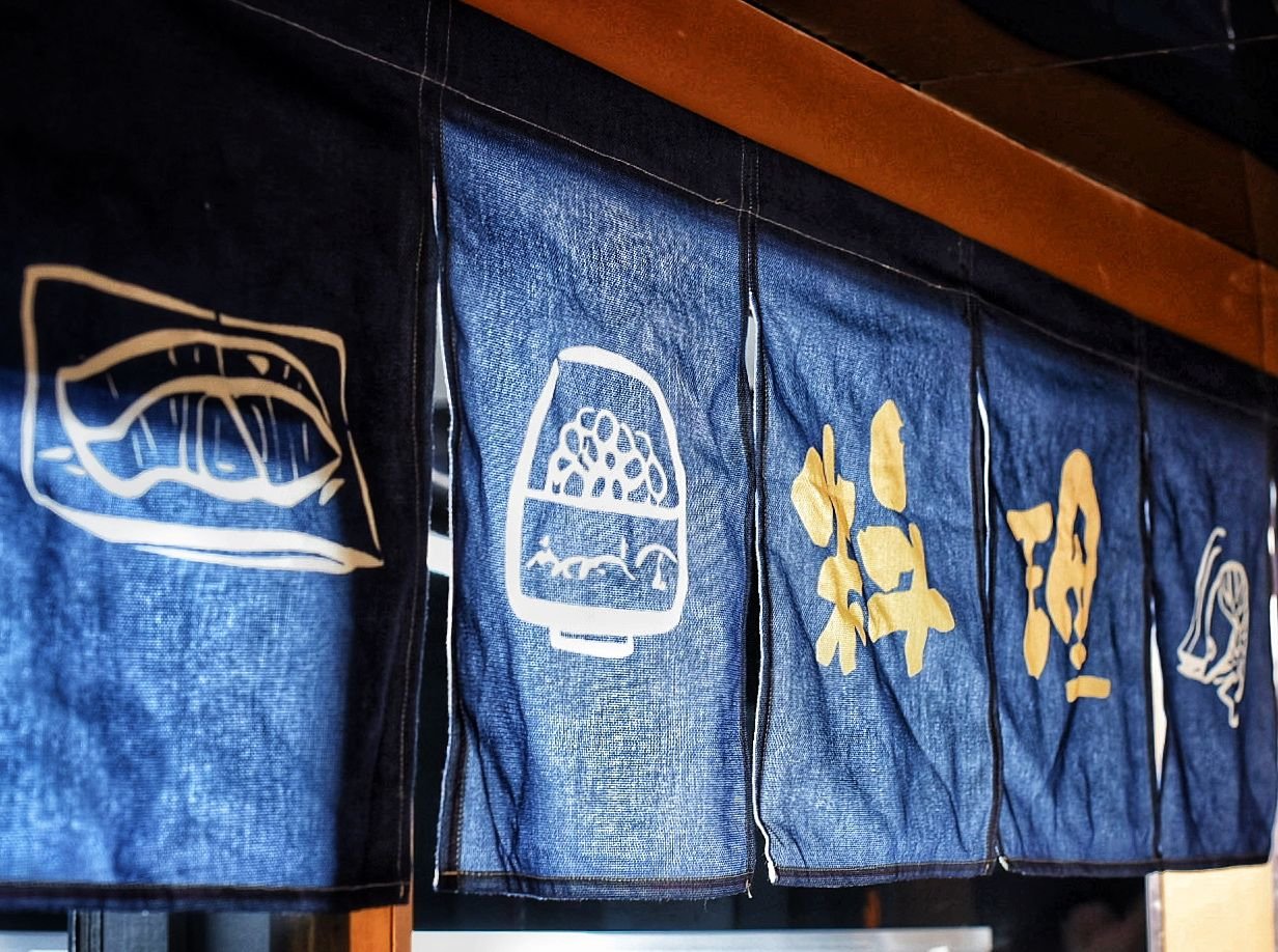 Japanese noren curtain with white and yellow symbols and illustrations related to food, hanging in front of a counter or doorway.