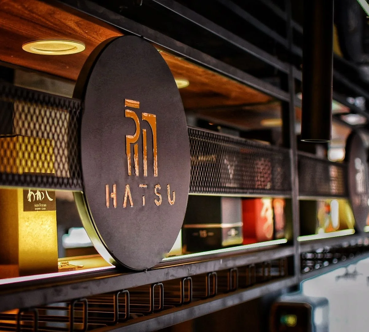 A black circular sign with orange cutout lettering and symbols, displaying the word "HATSÜ" and a stylized logo, mounted on a metal shelf with various boxes and items in the background.