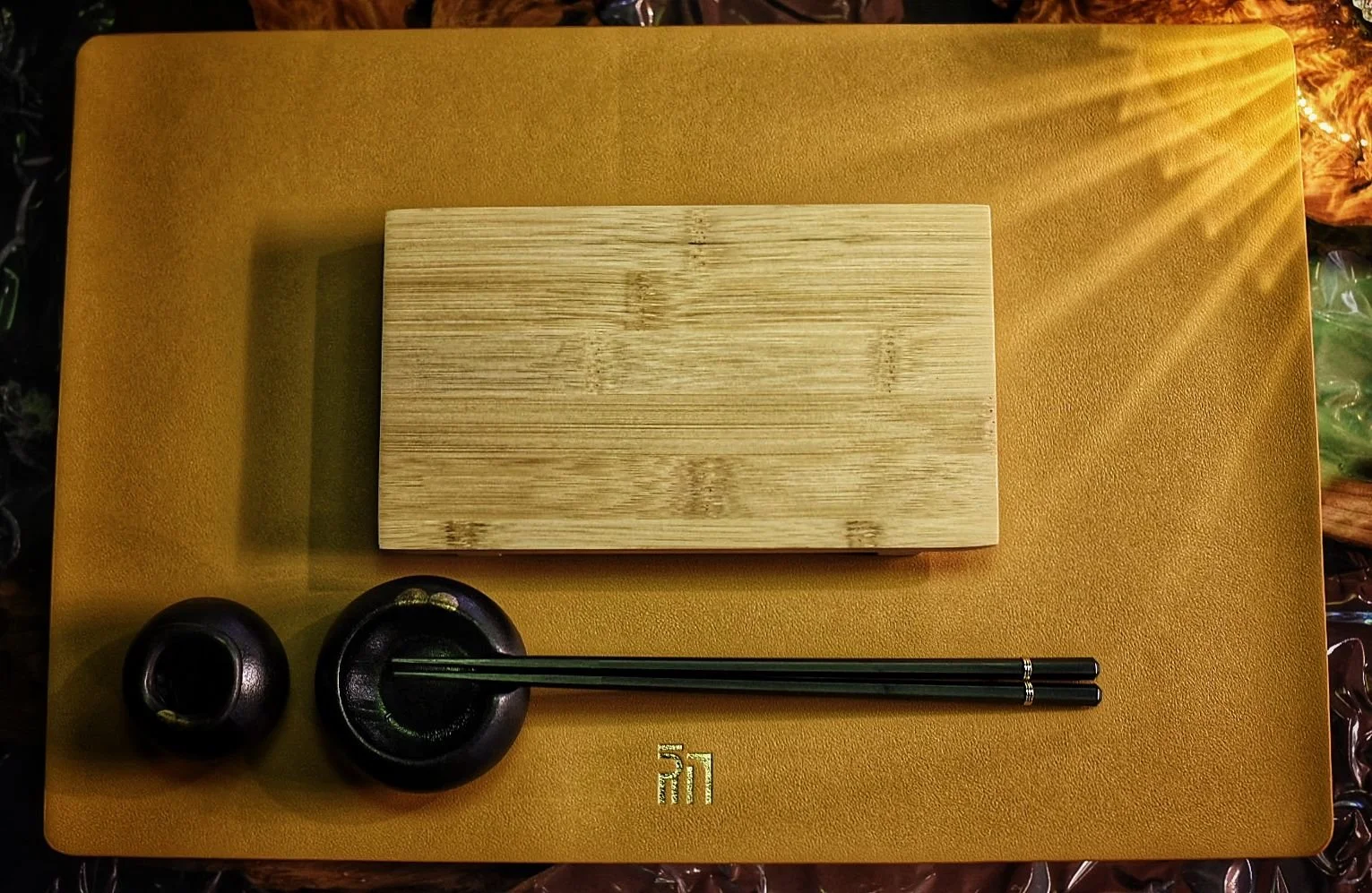 A wooden cutting board, a pair of black chopsticks, and two small black ceramic dishes are arranged on a yellow textured surface, which has a word in a foreign language written on it.