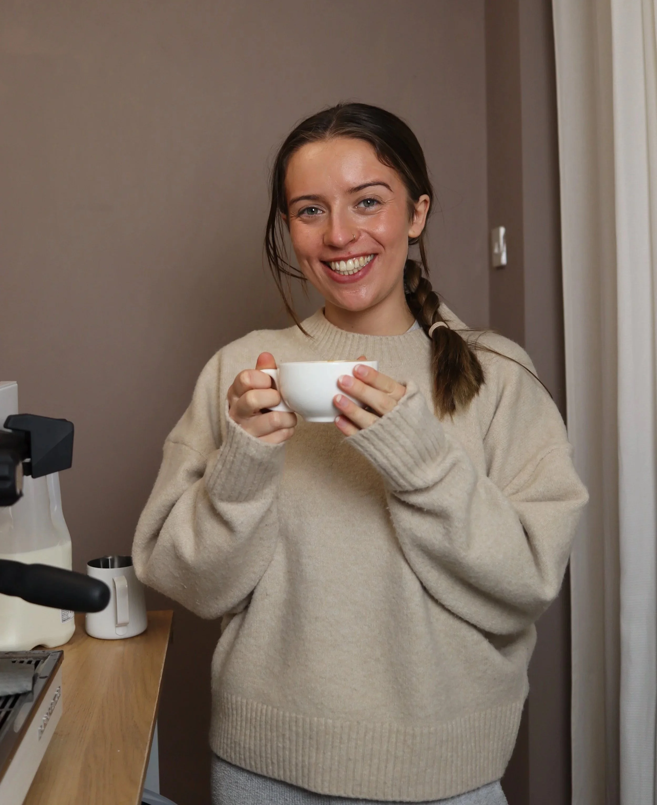 A young woman with a plait smiling and holding a white mug in what appears to be a kitchen setting.