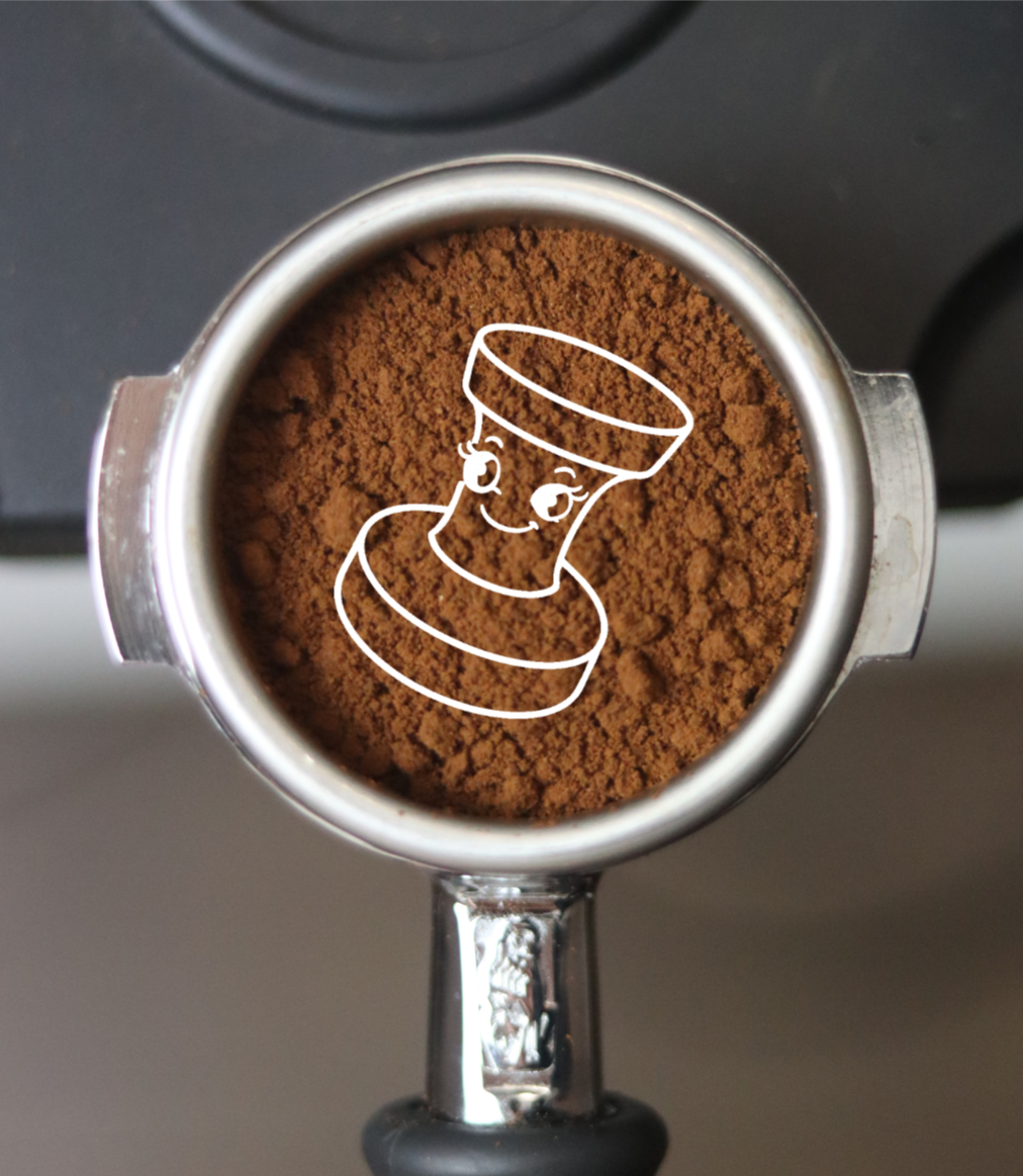 Top view of a portafilter filled with ground coffee, with a cute illustration of a smiling coffee cup with eyes and earrings drawn on it.