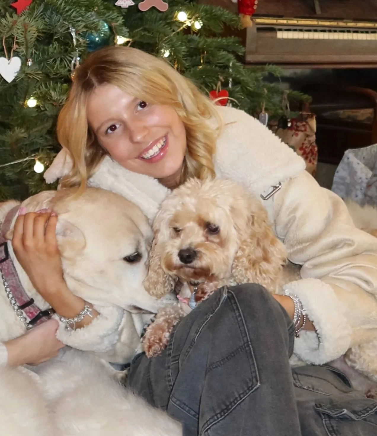 Woman with blonde hair smiling, surrounded by two dogs, in front of a decorated Christmas tree with ornaments and lights.