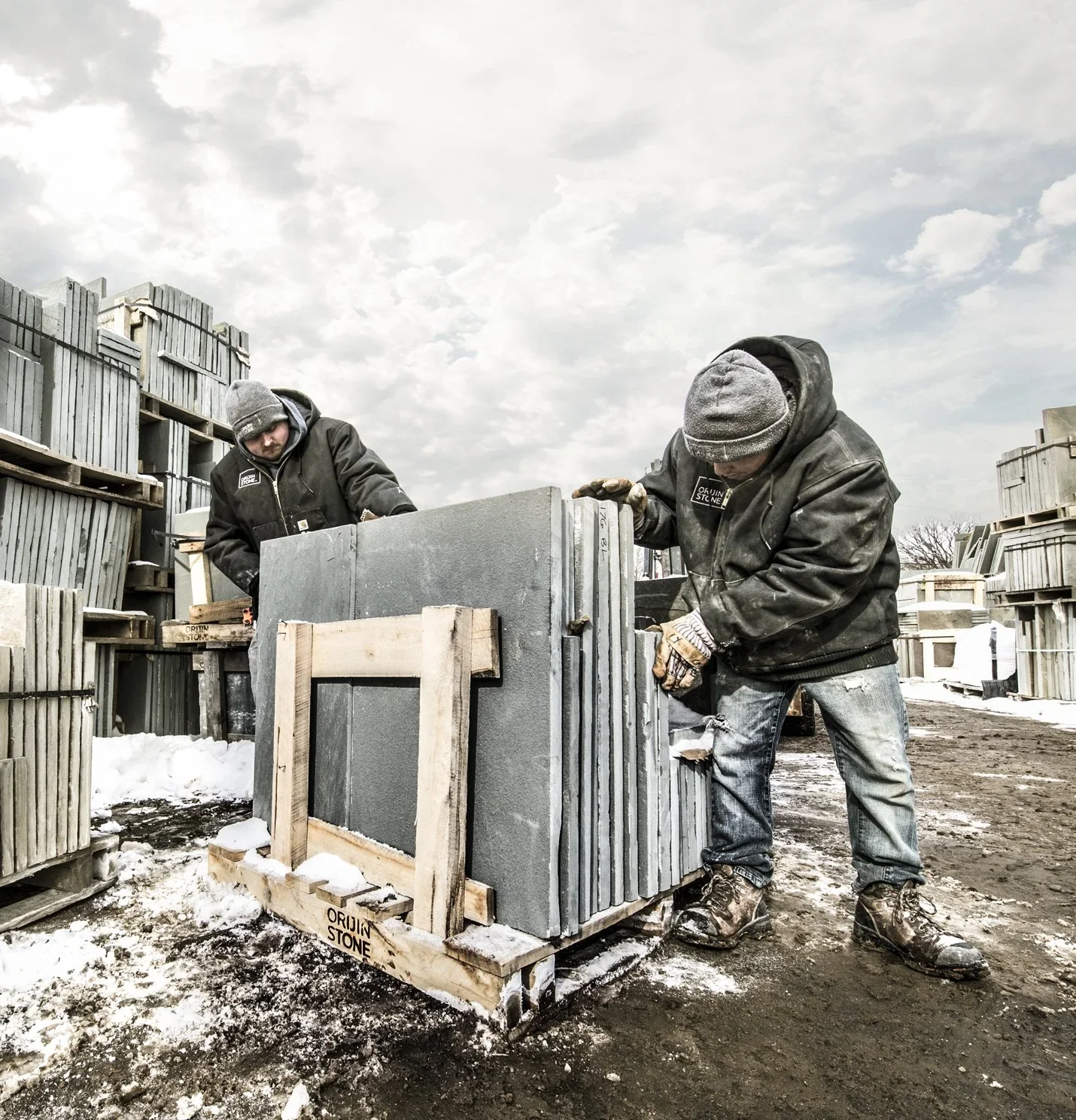 Two workers in winter clothing, including hats and gloves, are handling concrete slabs at an outdoor stone yard, with stacks of stone blocks around them and snow on the ground.