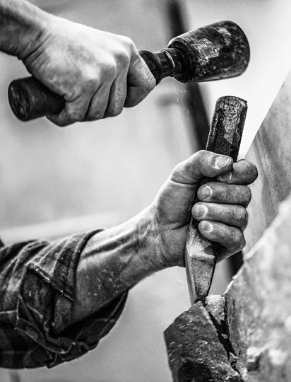 Close-up of two hands working with chisels and a hammer on a piece of stone or wood.