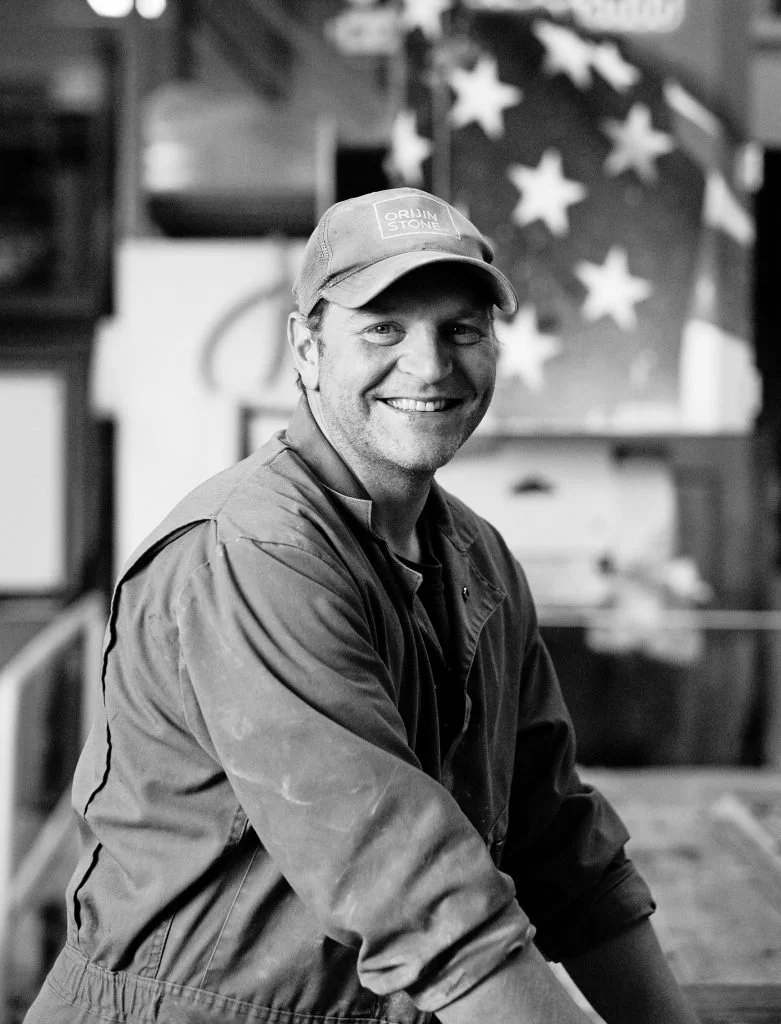 smiling man wearing a cap and jacket, with a blurred American flag in the background