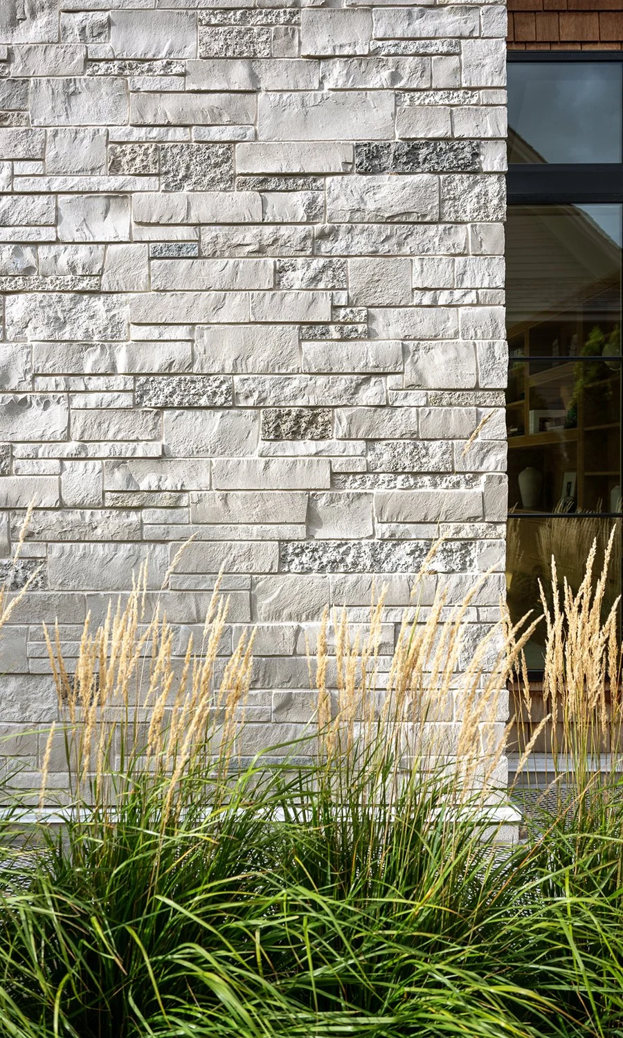 Close-up of a white stone brick wall with a window to the right, partially visible, and tall ornamental grass with beige seed heads in the foreground.