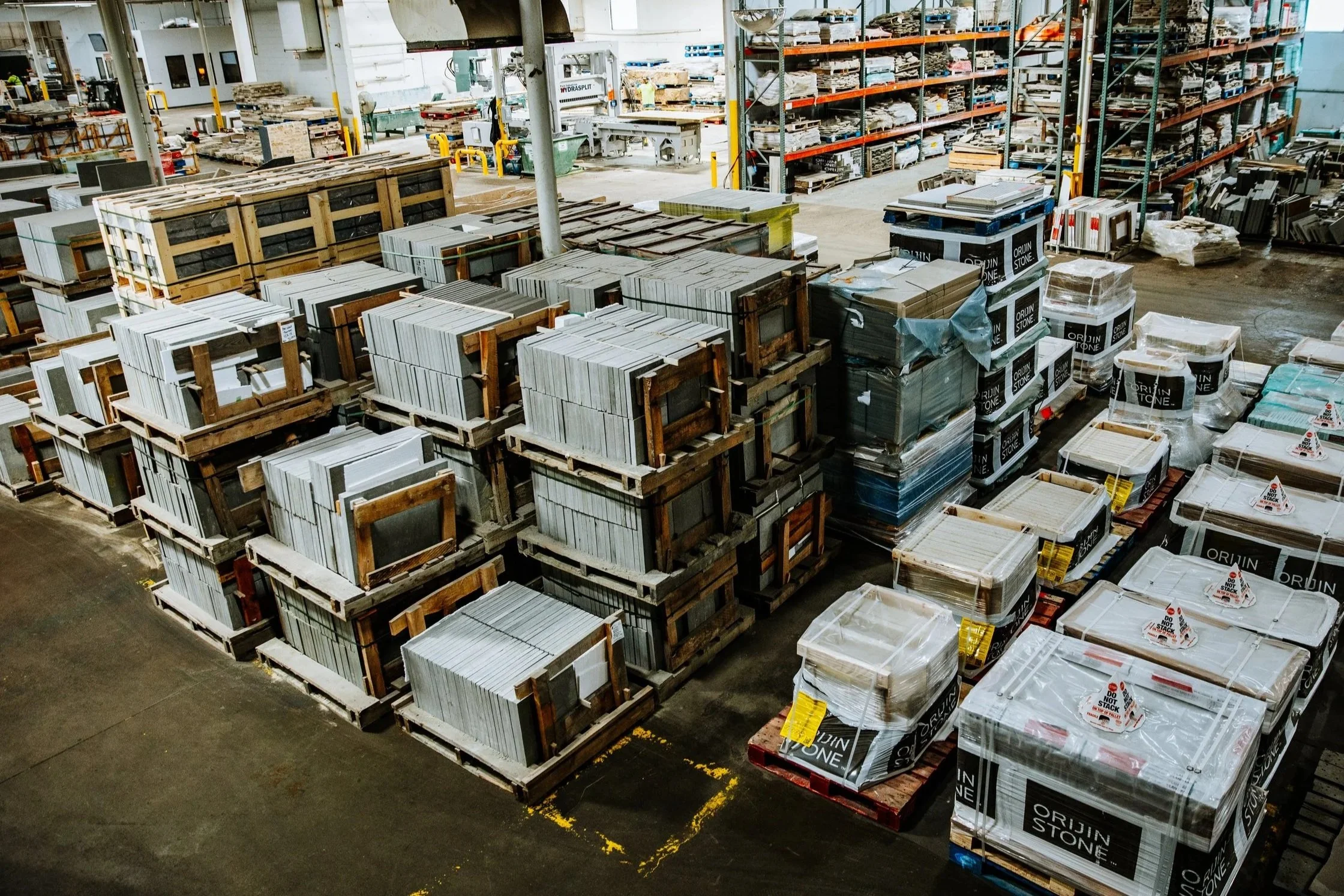 Warehouse interior with stacked tiles and stone slabs on pallets, shelves filled with materials, industrial equipment, and packaging supplies.