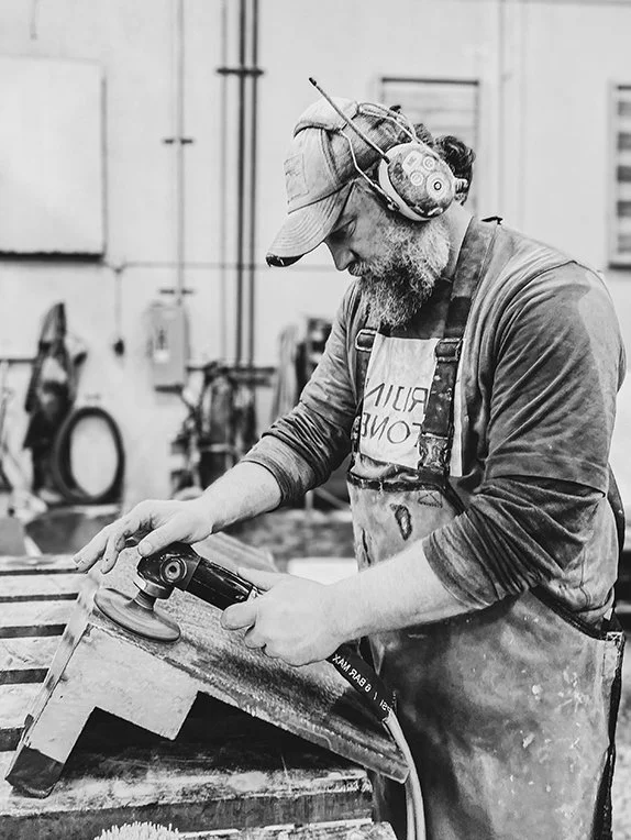 A man with a beard and glasses wearing a cap, headphones, and apron, uses a power sander on wood in a workshop.