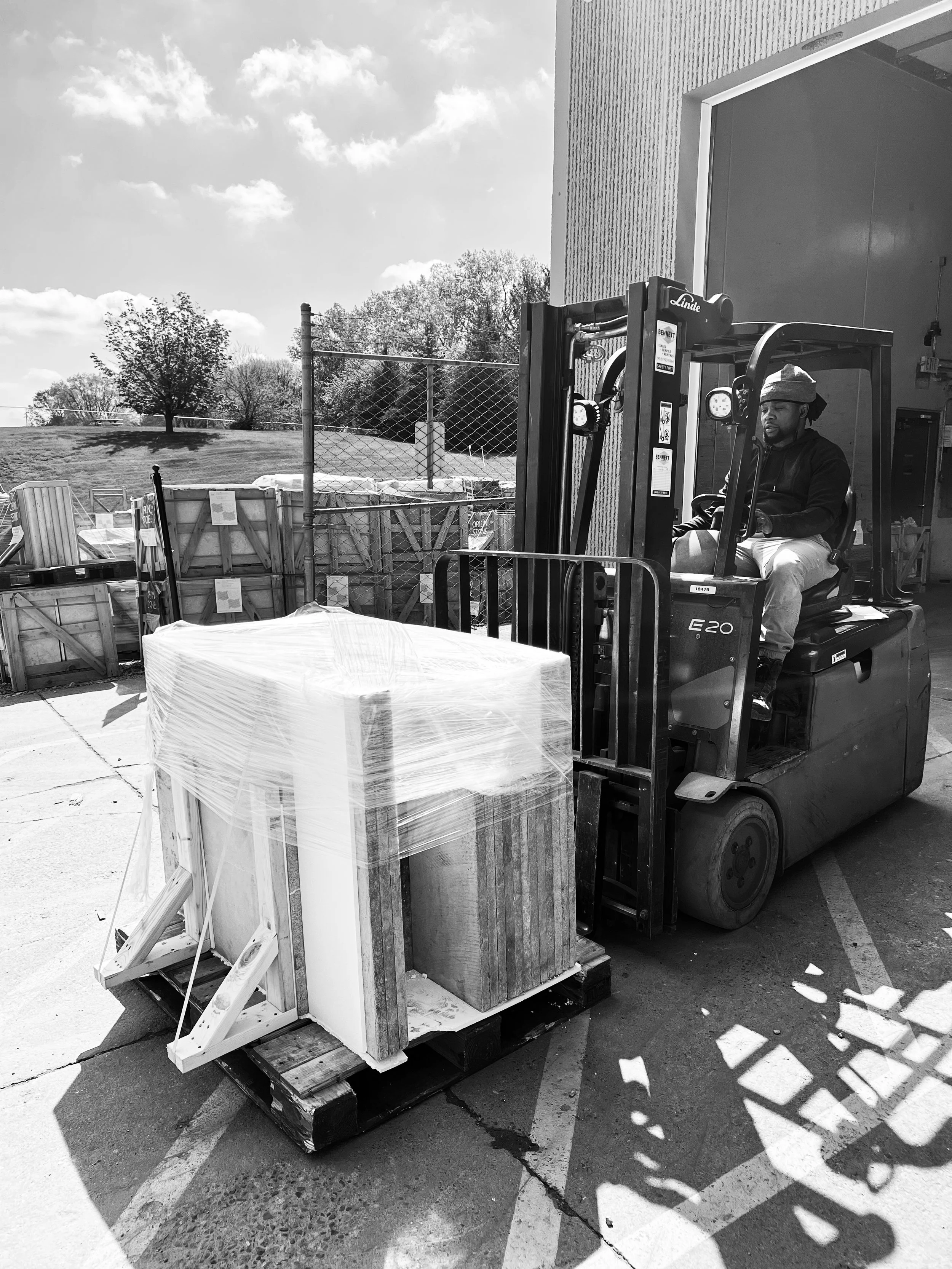 A man operating a forklift in an outdoor parking lot, lifting wooden crates wrapped in plastic around large rectangular objects. The scene features a partly cloudy sky, trees, and a chain-link fence in the background.
