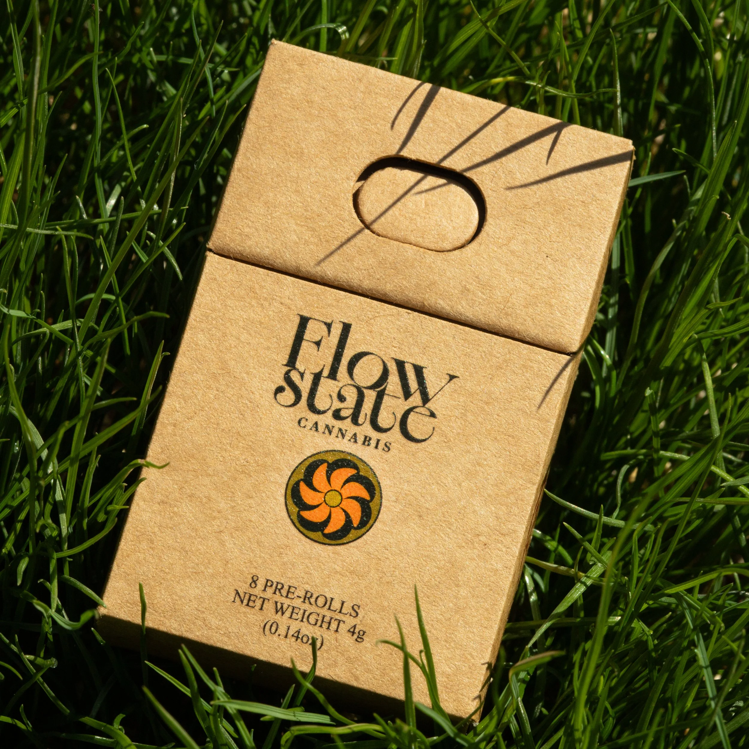 A cardboard pack of cannabis pre-rolls lying on green grass, with sunlight casting shadows from nearby blades.