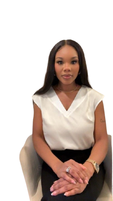 A woman with long dark hair, wearing a white top and black pants, sitting with hands clasped on her lap, against a plain background.