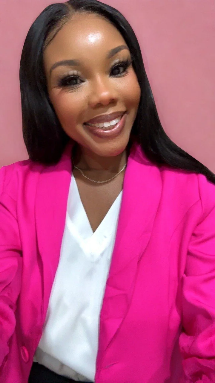 A woman smiling, wearing a bright pink blazer over a white blouse, has long black hair, and is posing against a pink background.