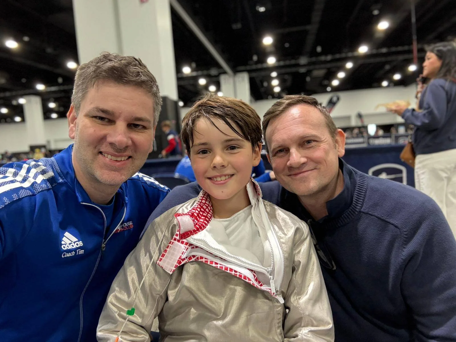 Two men and a boy smiling for a selfie at an indoor sports venue. The boy is wearing a racing suit, and the men are dressed casually, one in a blue Adidas jacket and the other in a dark sweater.