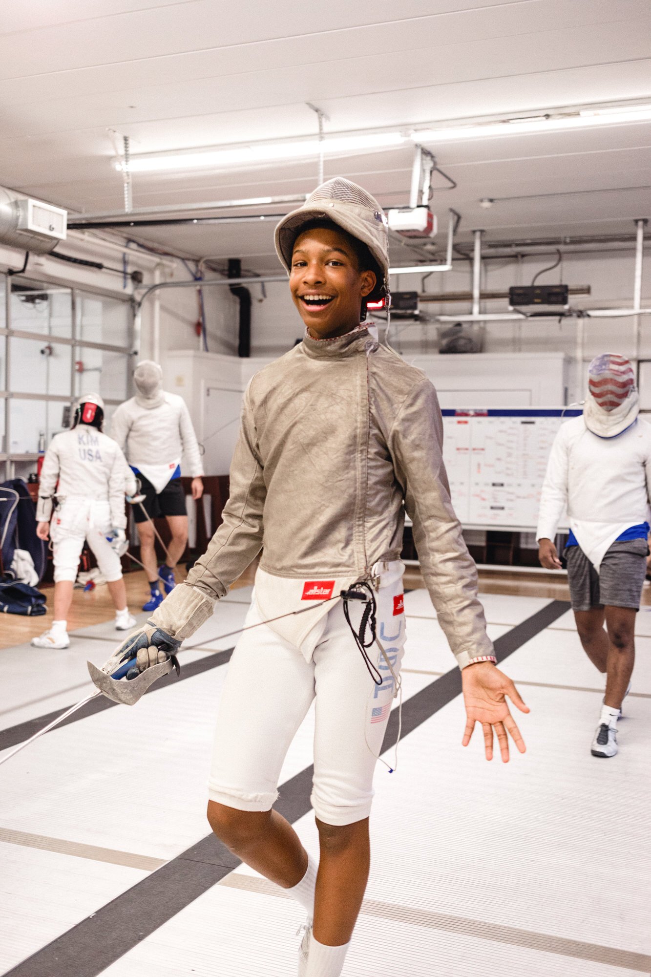 Young fencer smiling in a fencing gym, preparing for practice or competition, with other fencers in the background.