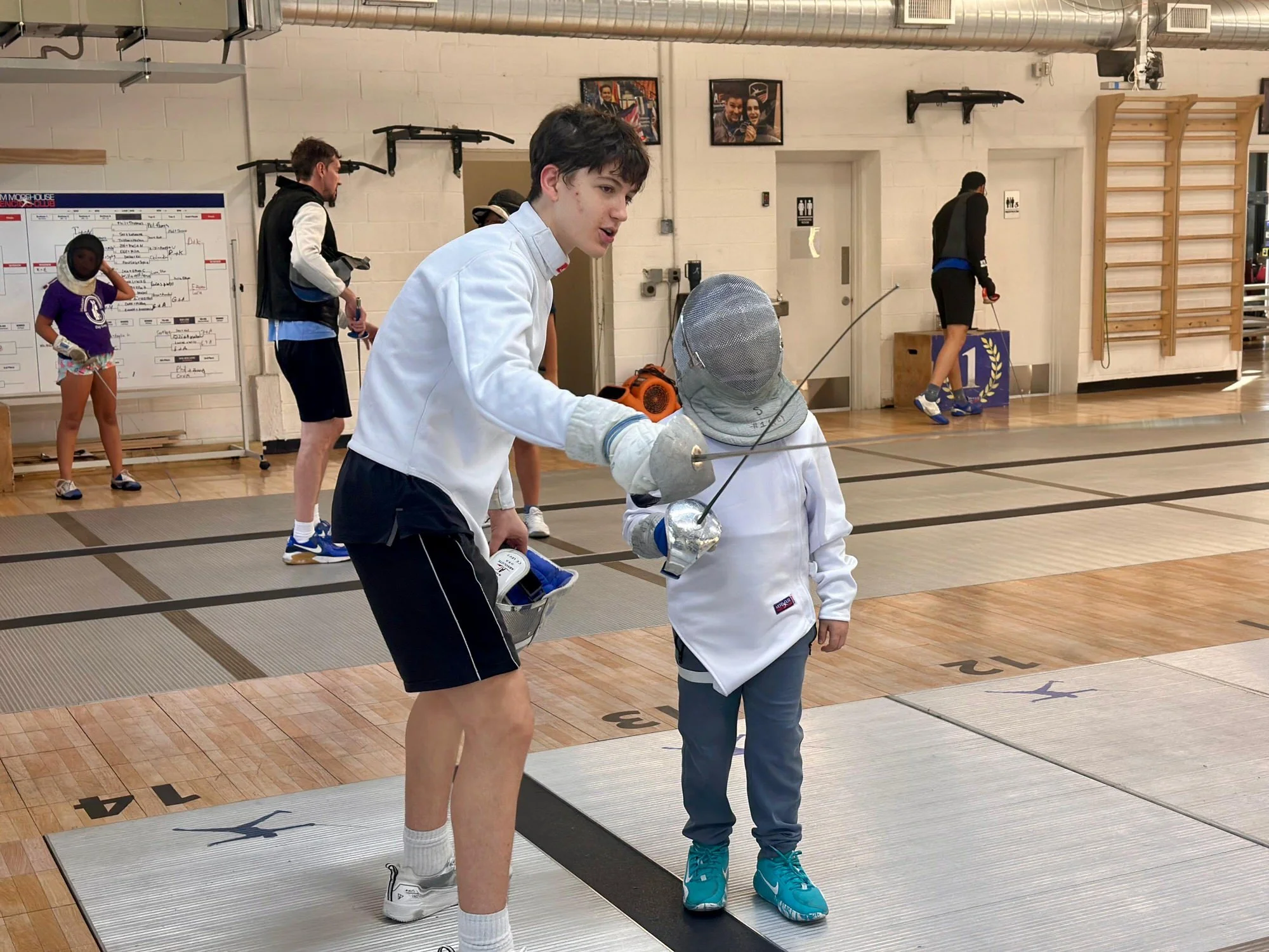 Young male fencing instructor teaches fencing technique to a young girl in fencing gear at an indoor training facility.